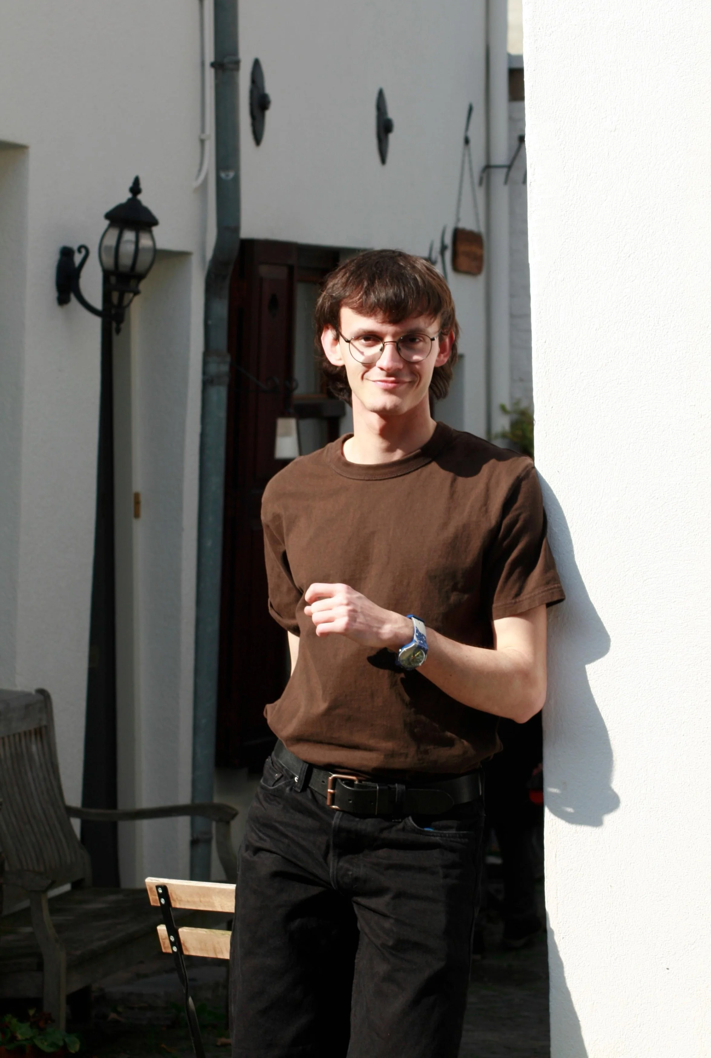 A young man with brown hair, glasses, wearing a brown shirt and black pants, standing outdoors in front of a white wall and smiling.