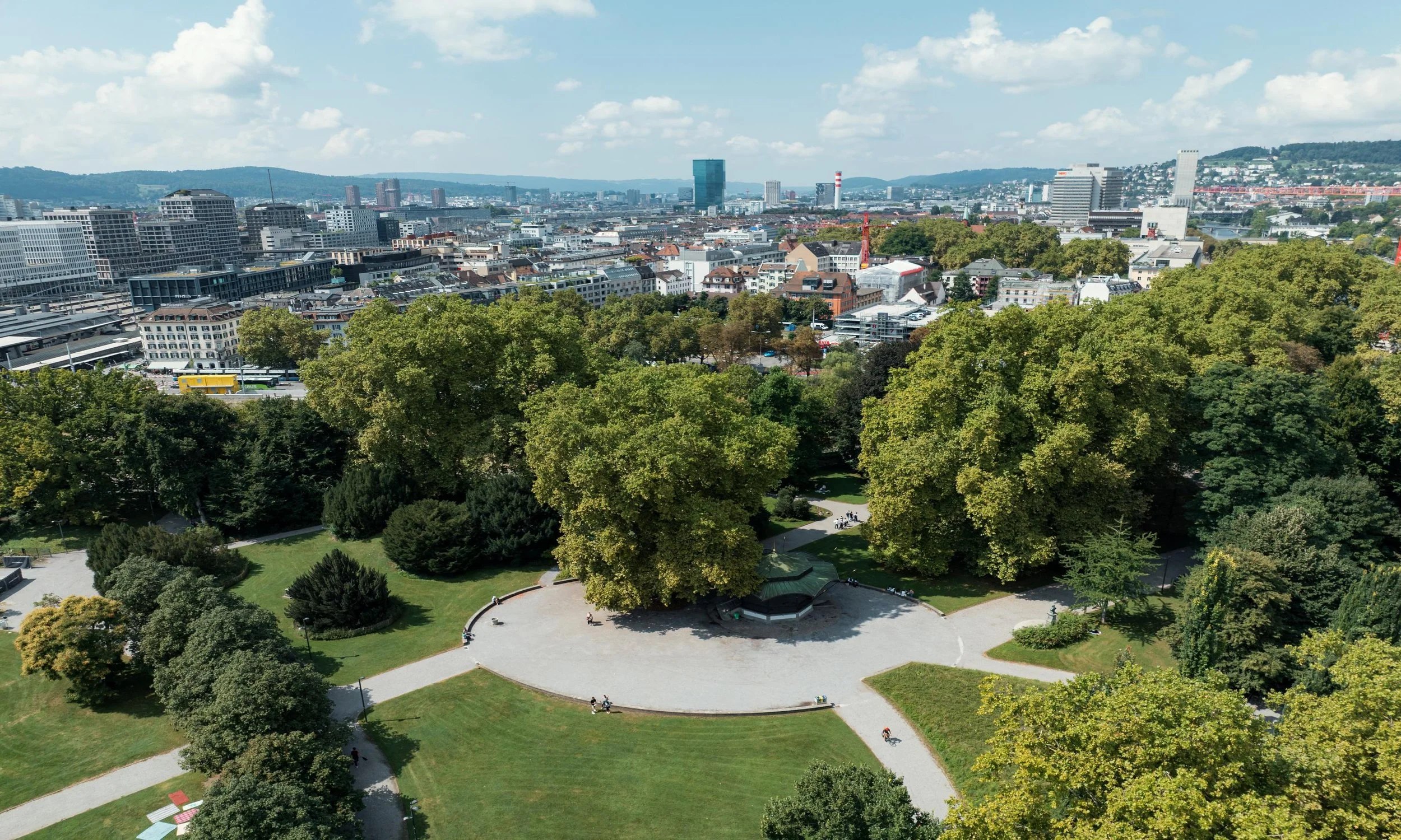 A city skyline with modern high-rise buildings in the background, and a park with lush green trees and a circular walking path in the foreground.