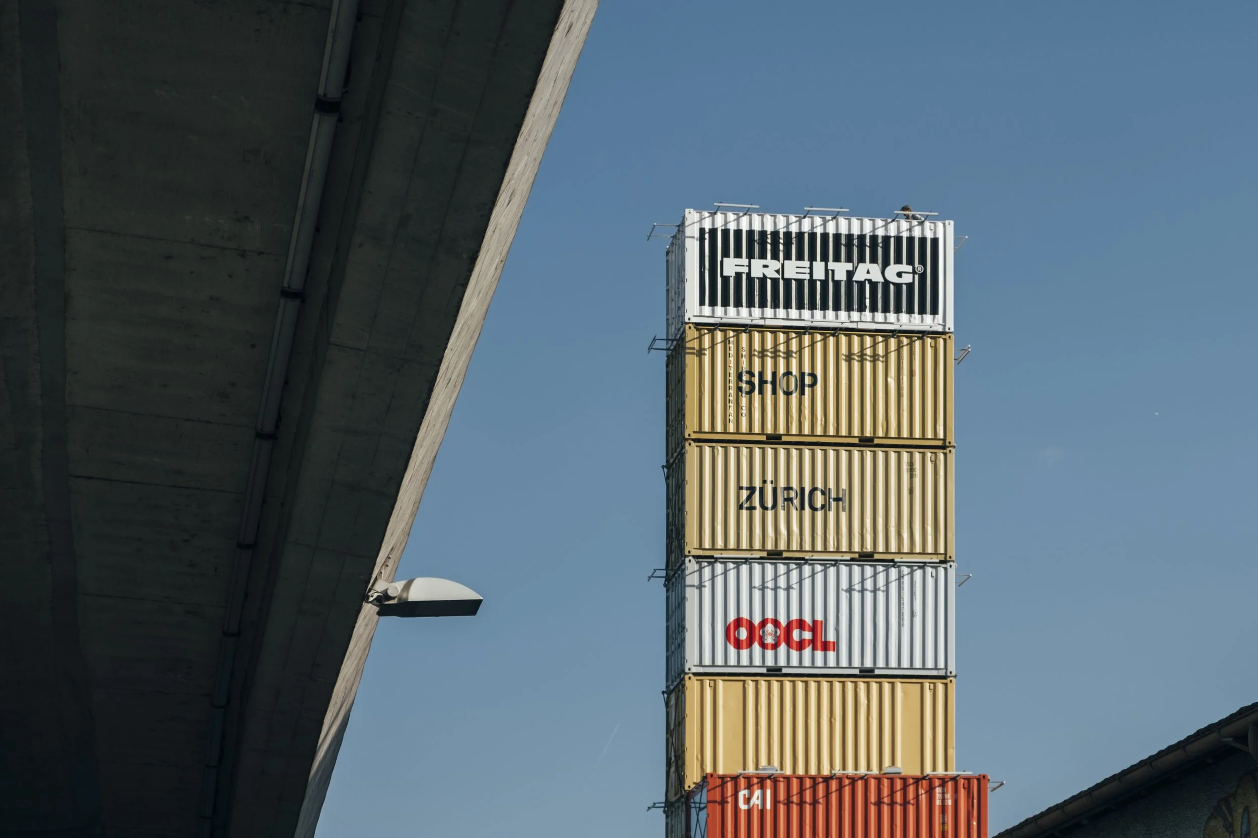 Stack of shipping containers beneath a bridge with a clear blue sky.