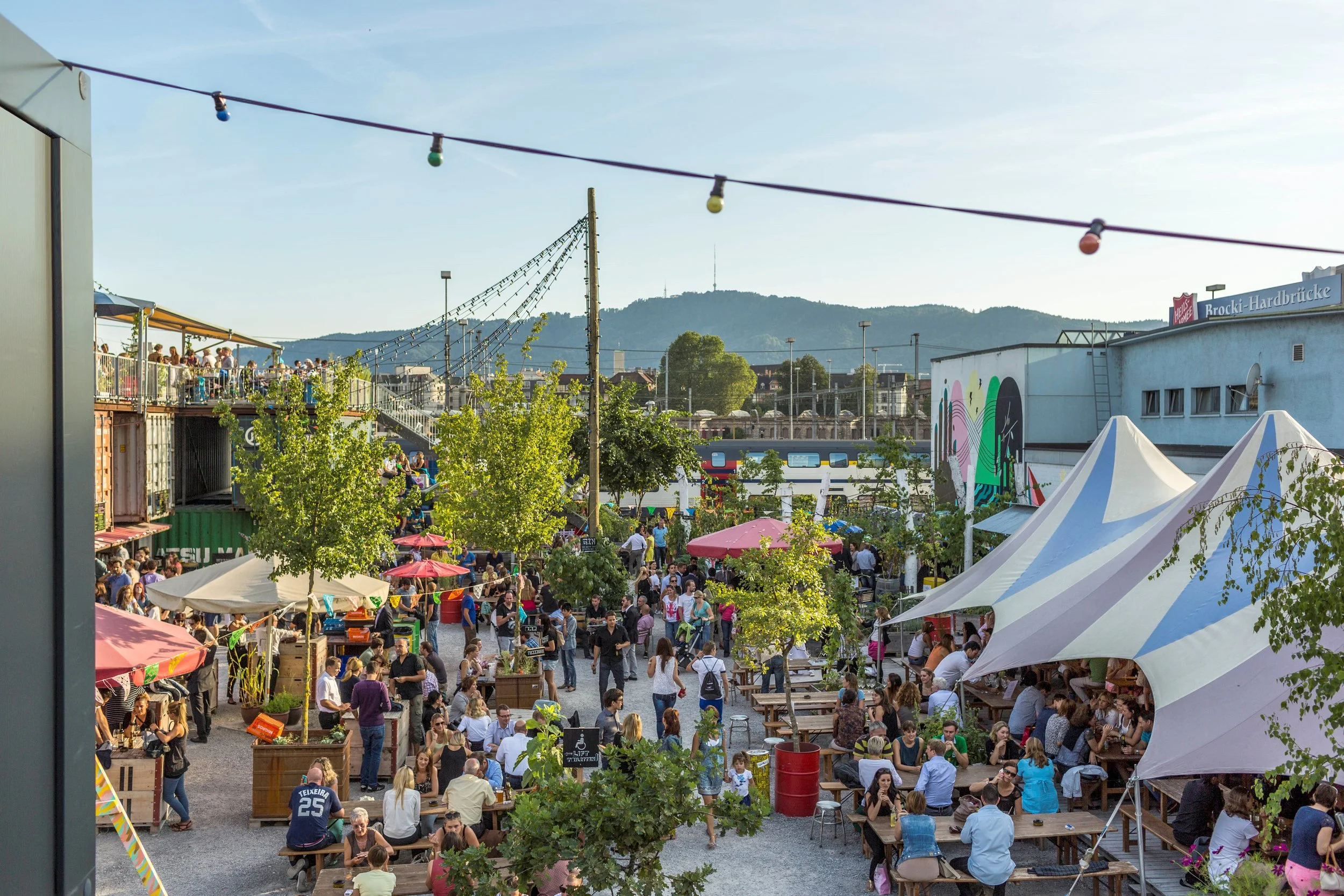 A lively outdoor market or event with many people mingling, sitting at tables, and walking around under white tents. Colorful umbrellas and string lights are visible, surrounded by greenery and city buildings with a mountain in the background.
