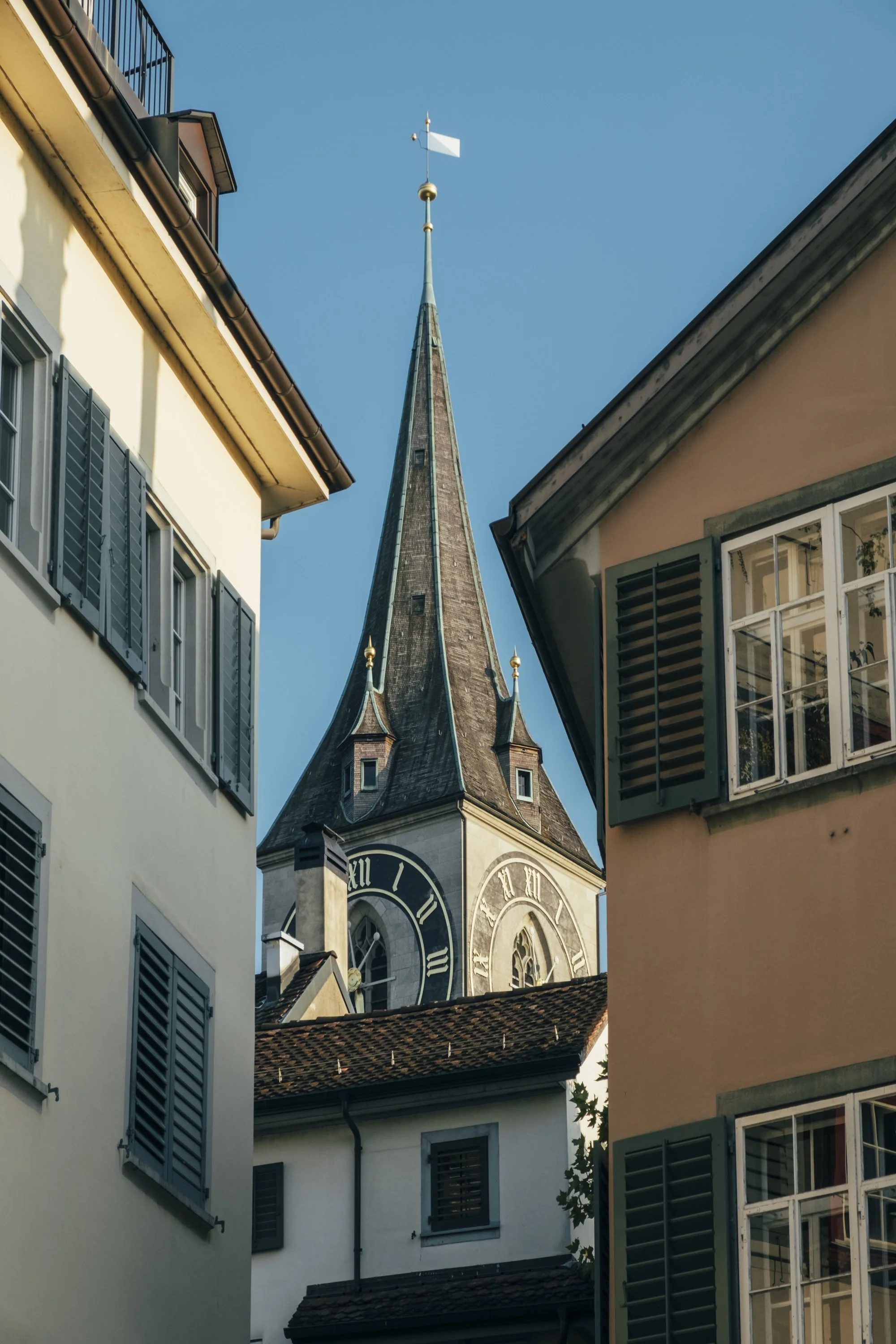 A church tower with a pointed steeple topped by a weather vane, framed by two residential buildings with windows and shutters, under a clear blue sky.