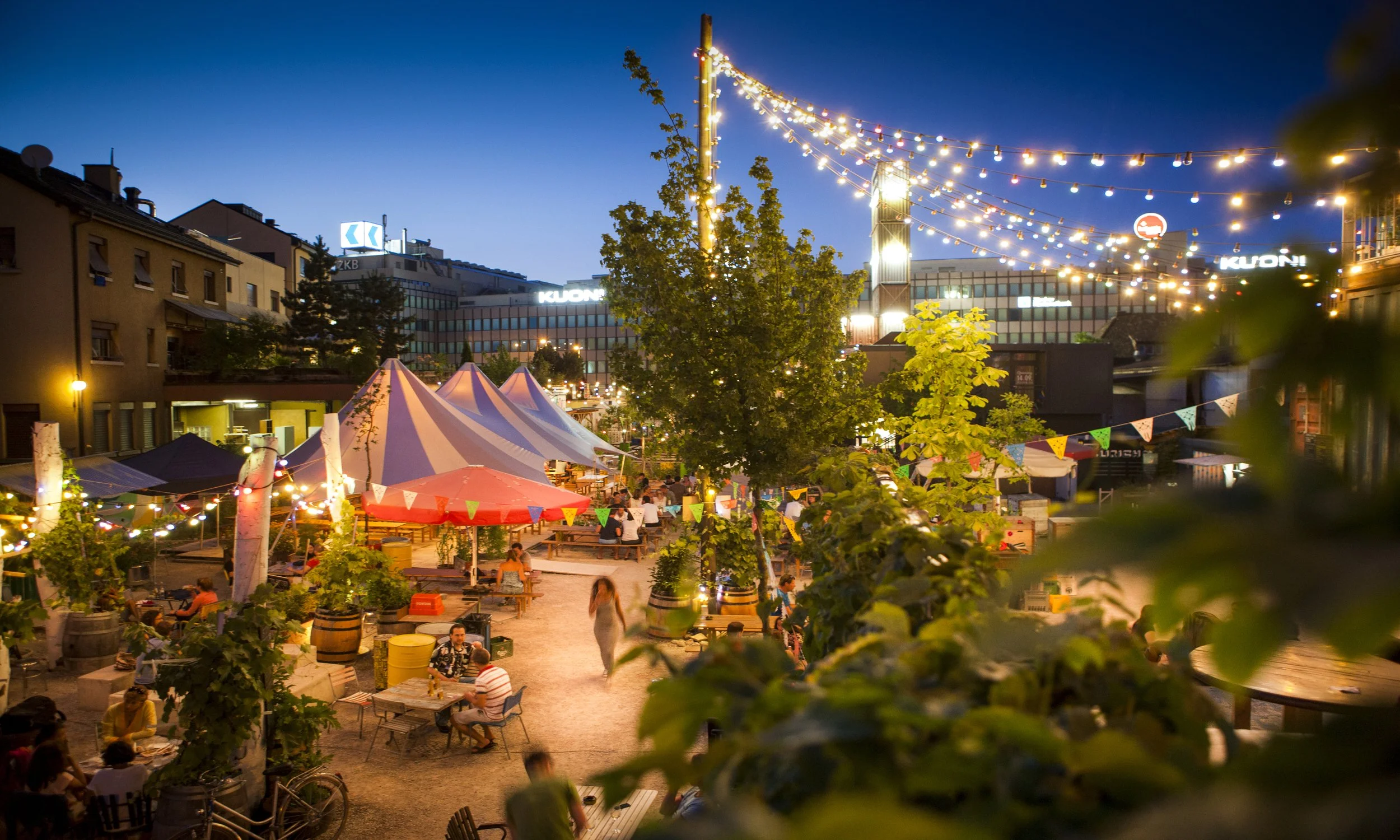 An outdoor evening scene with string lights, tents, benches, and people dining at tables in a lively urban setting, with buildings and trees in the background.