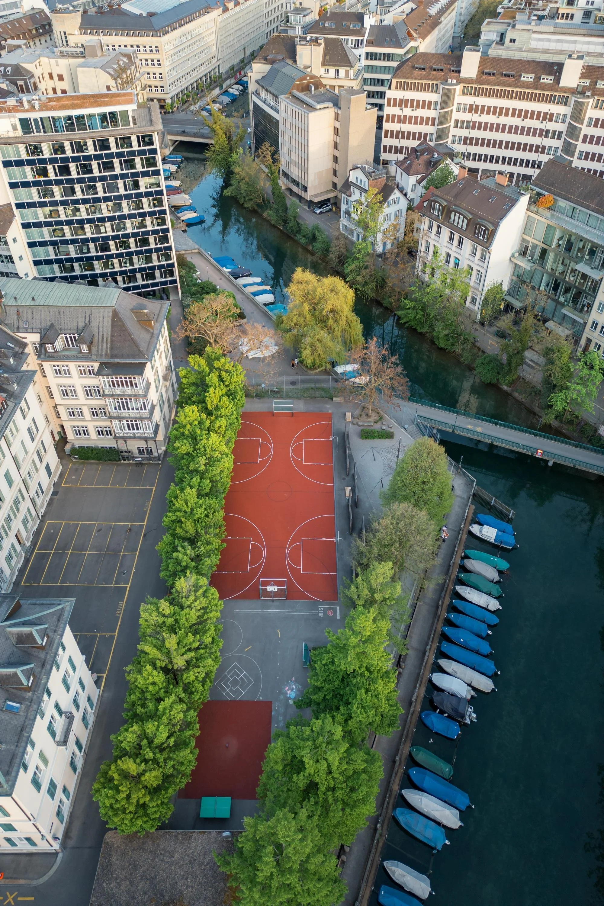 Aerial view of an urban area featuring a basketball court, a canal with boats along the quay, surrounded by tall buildings, trees, and parked cars.