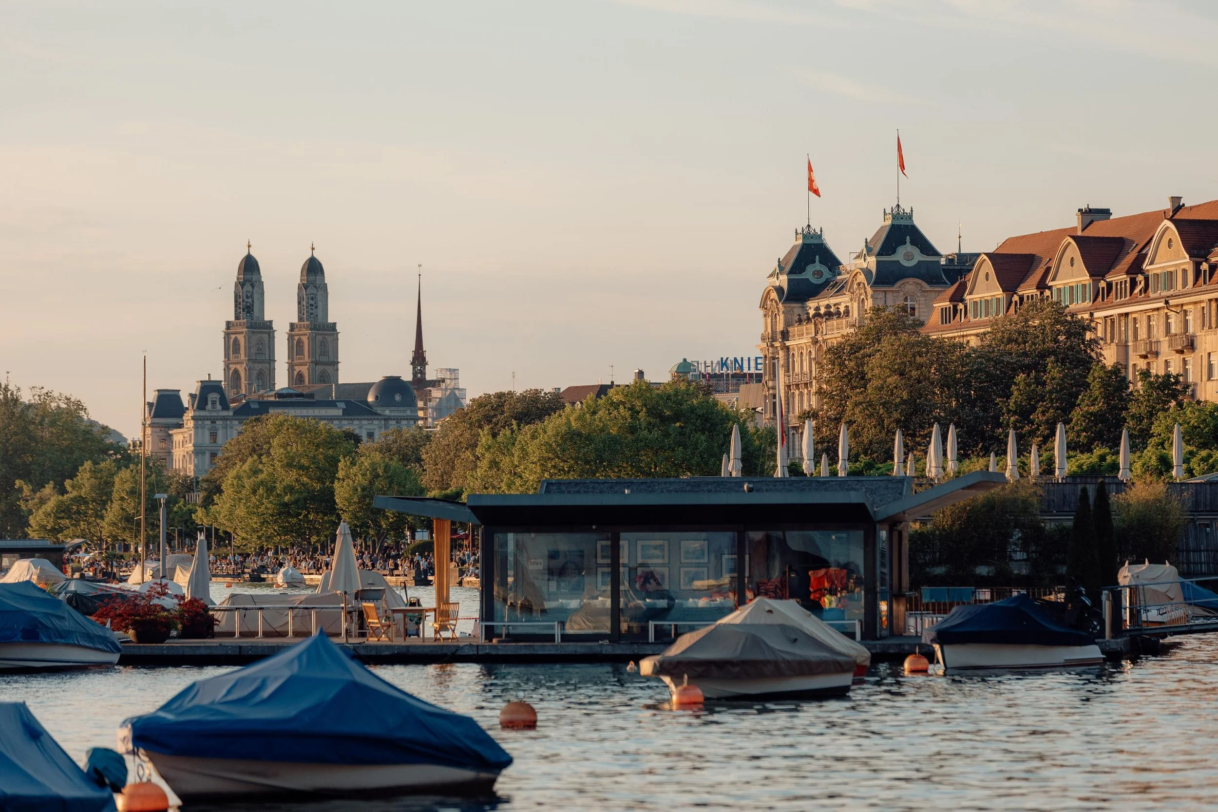 A scenic view of a city skyline with historic buildings, church spires, and flags, viewed over a marina with boats and umbrellas along the waterfront during late afternoon or early evening.