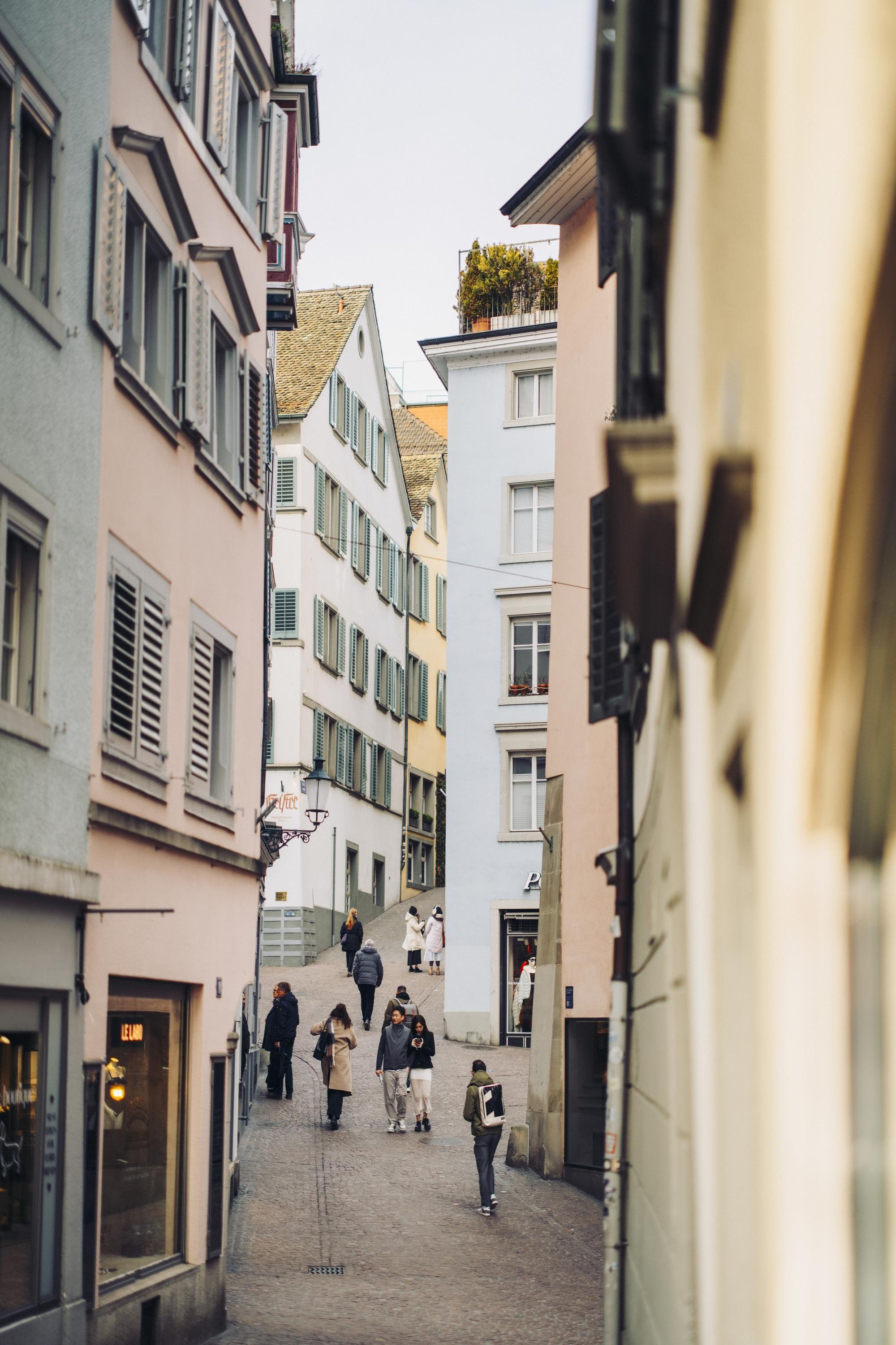 A narrow pedestrian street in a European city surrounded by pastel-colored buildings with shutters, with people walking and shopping.