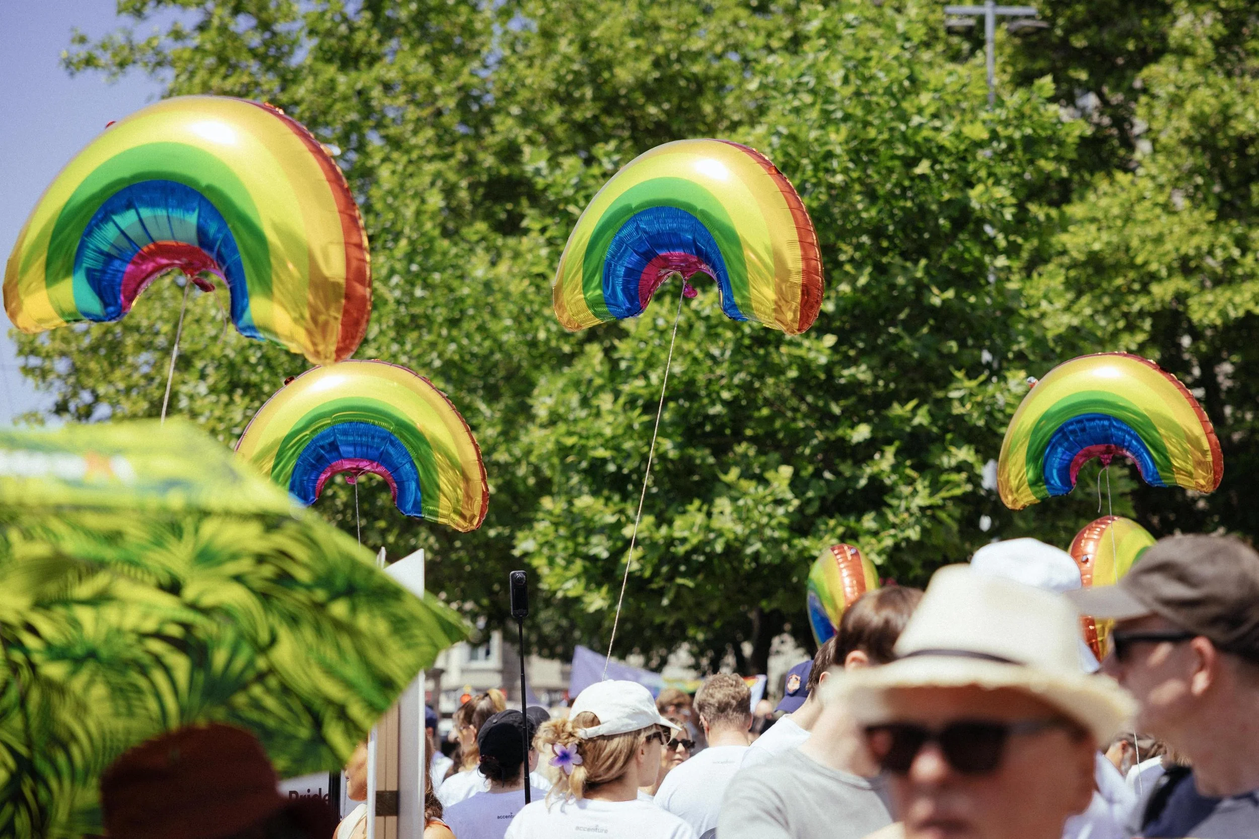 Colorful rainbow-shaped balloons floating above a crowd of people at an outdoor event, with green trees in the background.