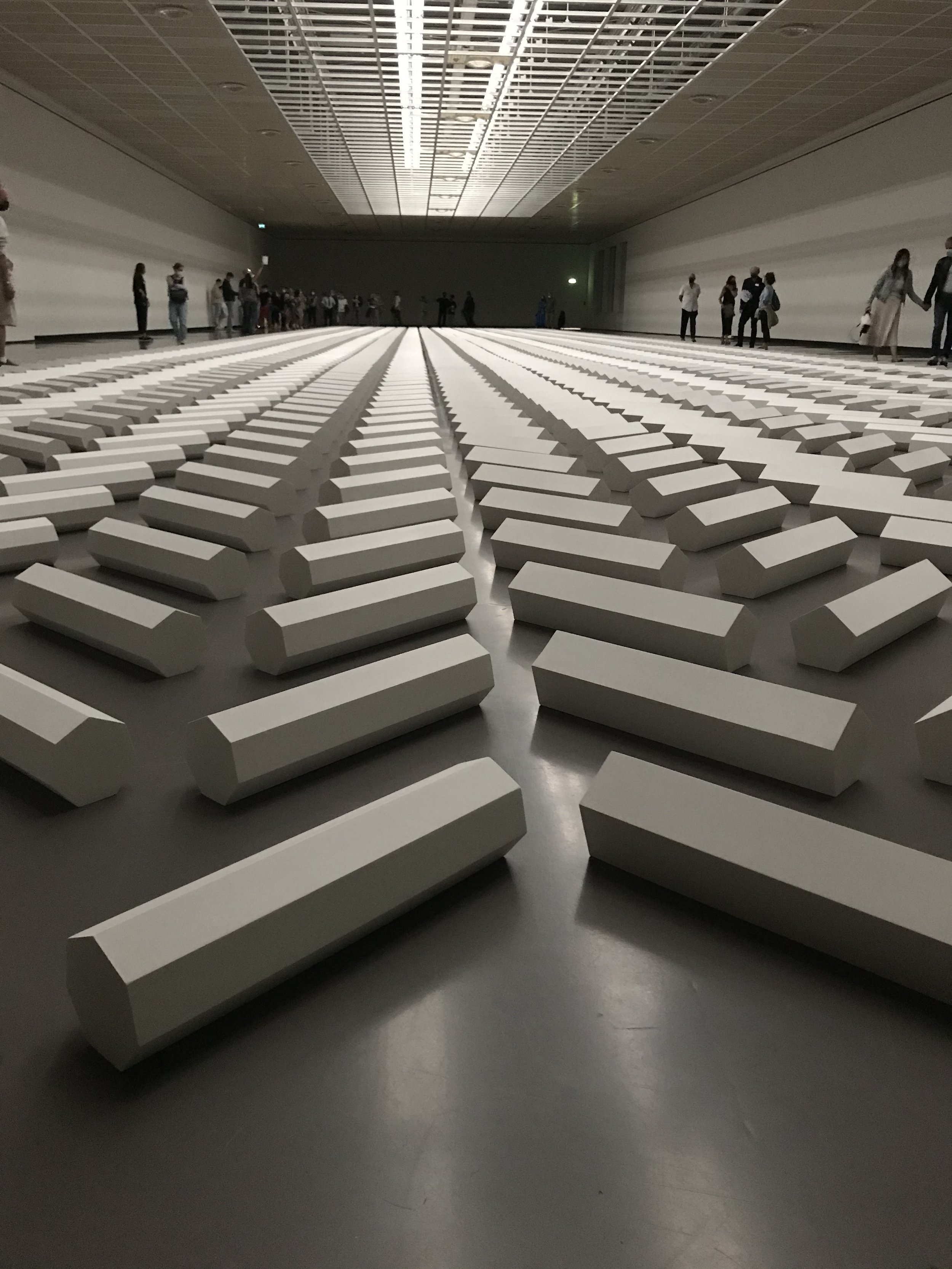 An indoor art installation consisting of numerous white rectangular blocks arranged in parallel rows on a flat surface. People are walking and observing in the background within a large, dimly lit gallery space with a high ceiling.