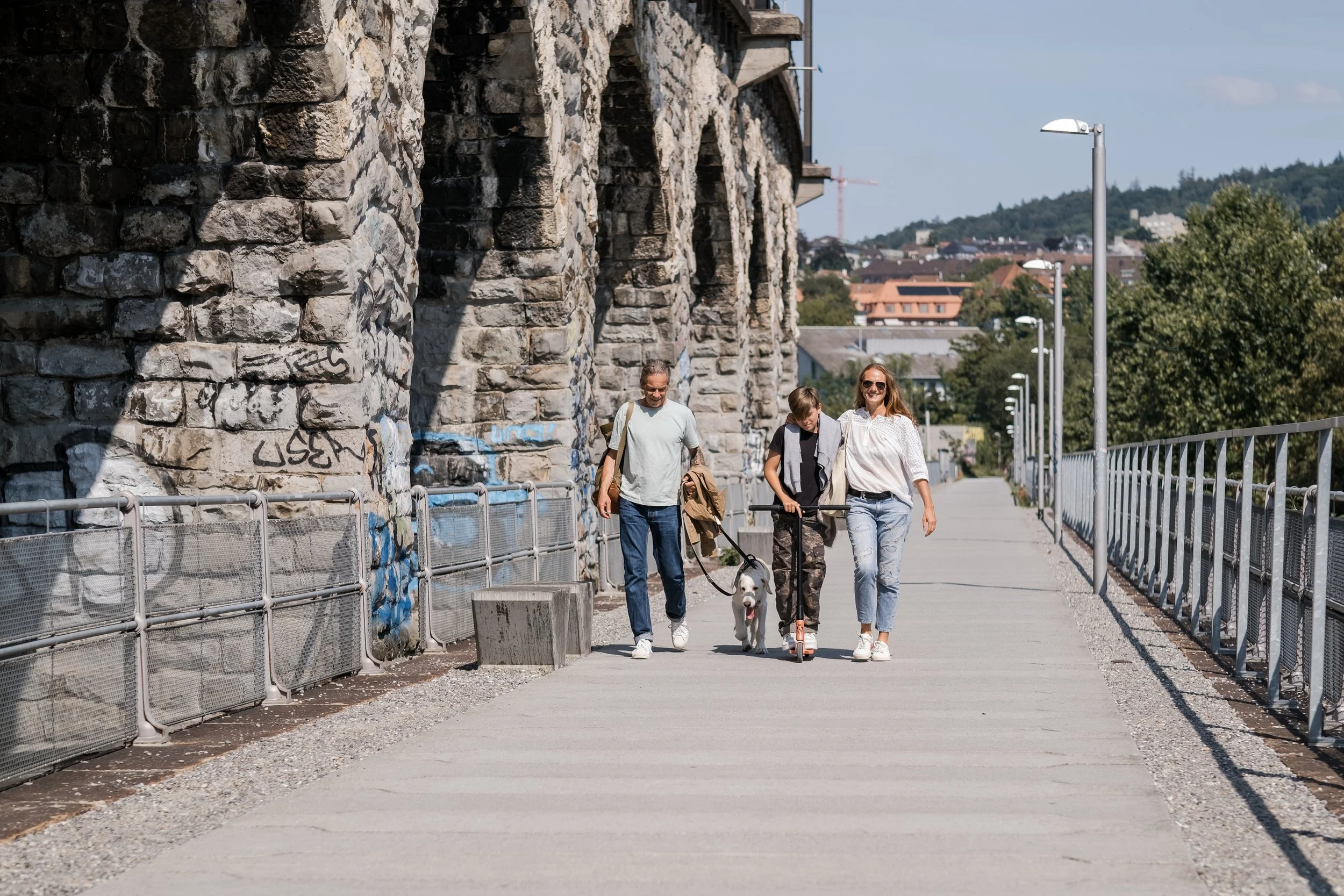 A family walking on a pedestrian bridge, with a stone wall on one side and street lamps along the pathway. The family includes a woman, a man, a boy walking with a dog, and a woman with sunglasses. The background shows houses and a hill with trees.