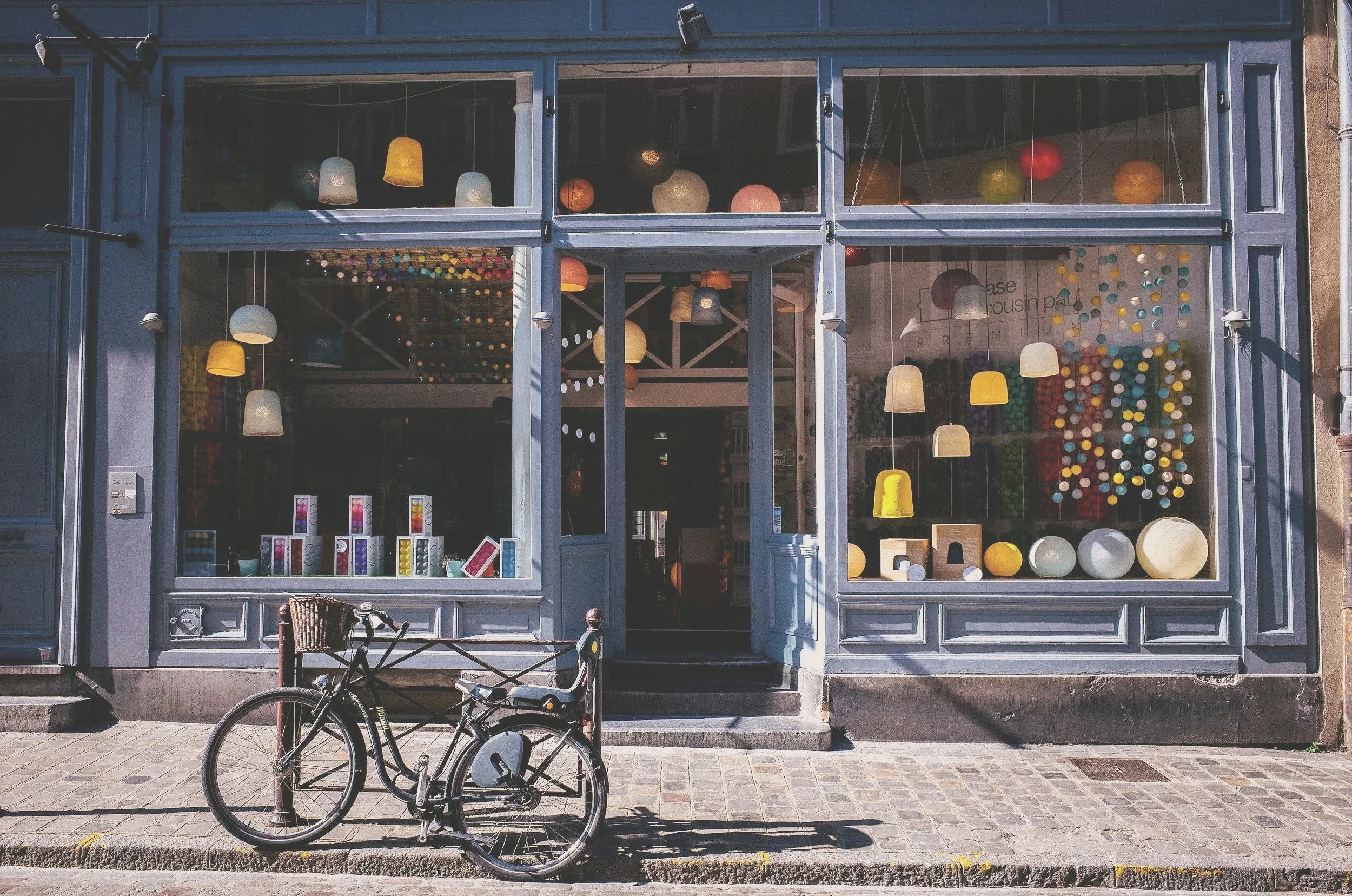 Small business storefront in Montreal with large glass windows displaying colorful lamps and a bicycle parked outside on cobblestone sidewalk.