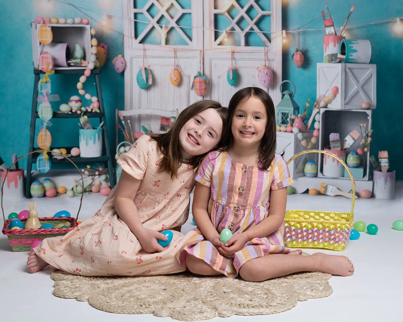 Two young girls sitting on a rug, holding plastic eggs, with Easter baskets and decorative decorations in the background.