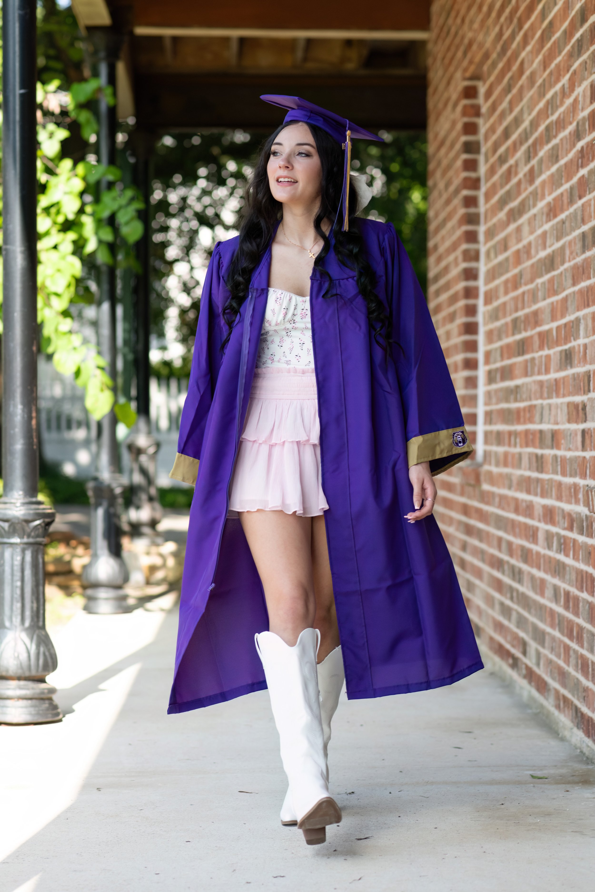 A young woman wearing a purple graduation cap and gown with a white dress underneath and white cowboy boots walking outdoors near a brick building.