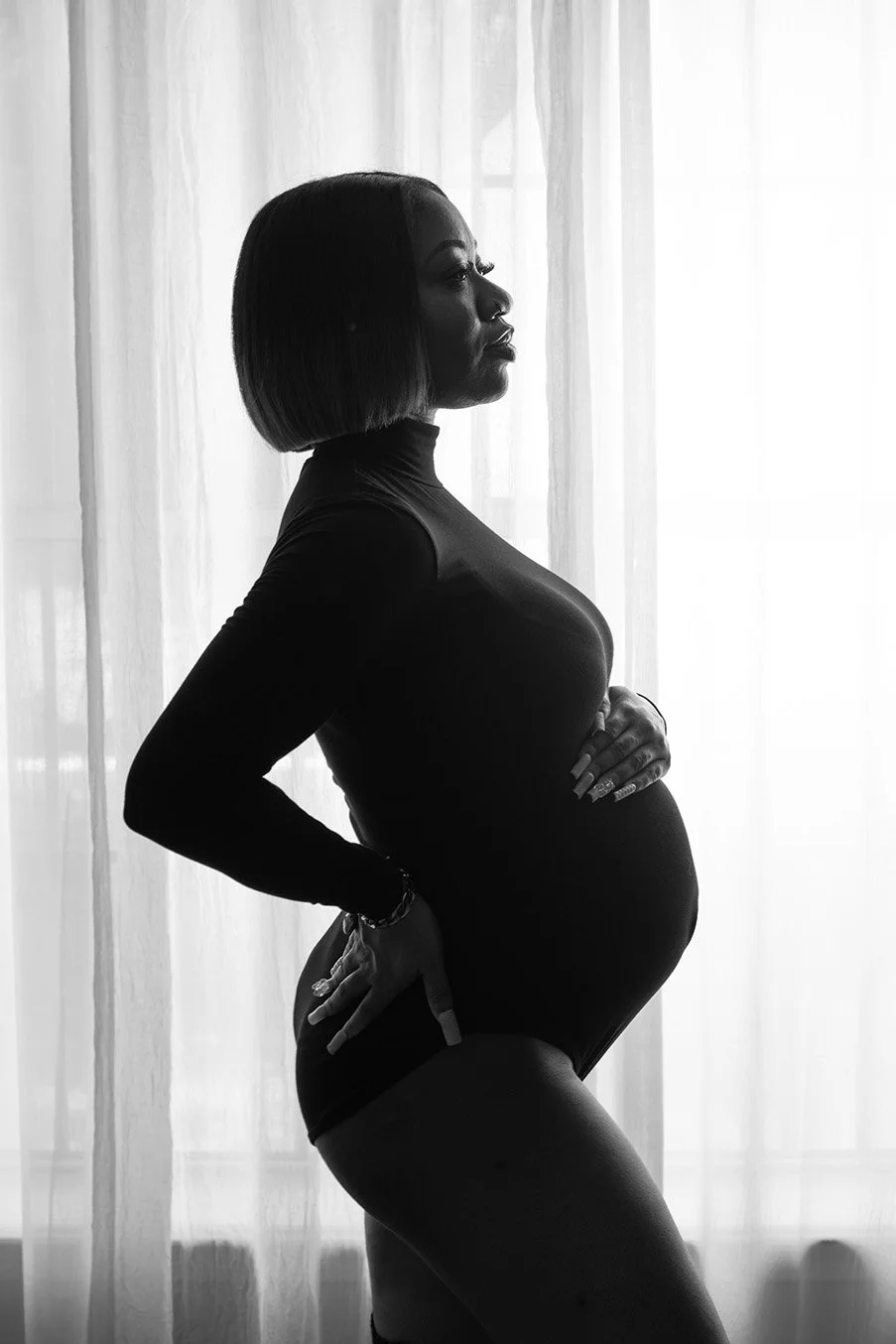 Black and white silhouette of a pregnant woman standing in profile in front of a sheer curtain, holding her belly with one hand and resting her other hand on her hip.