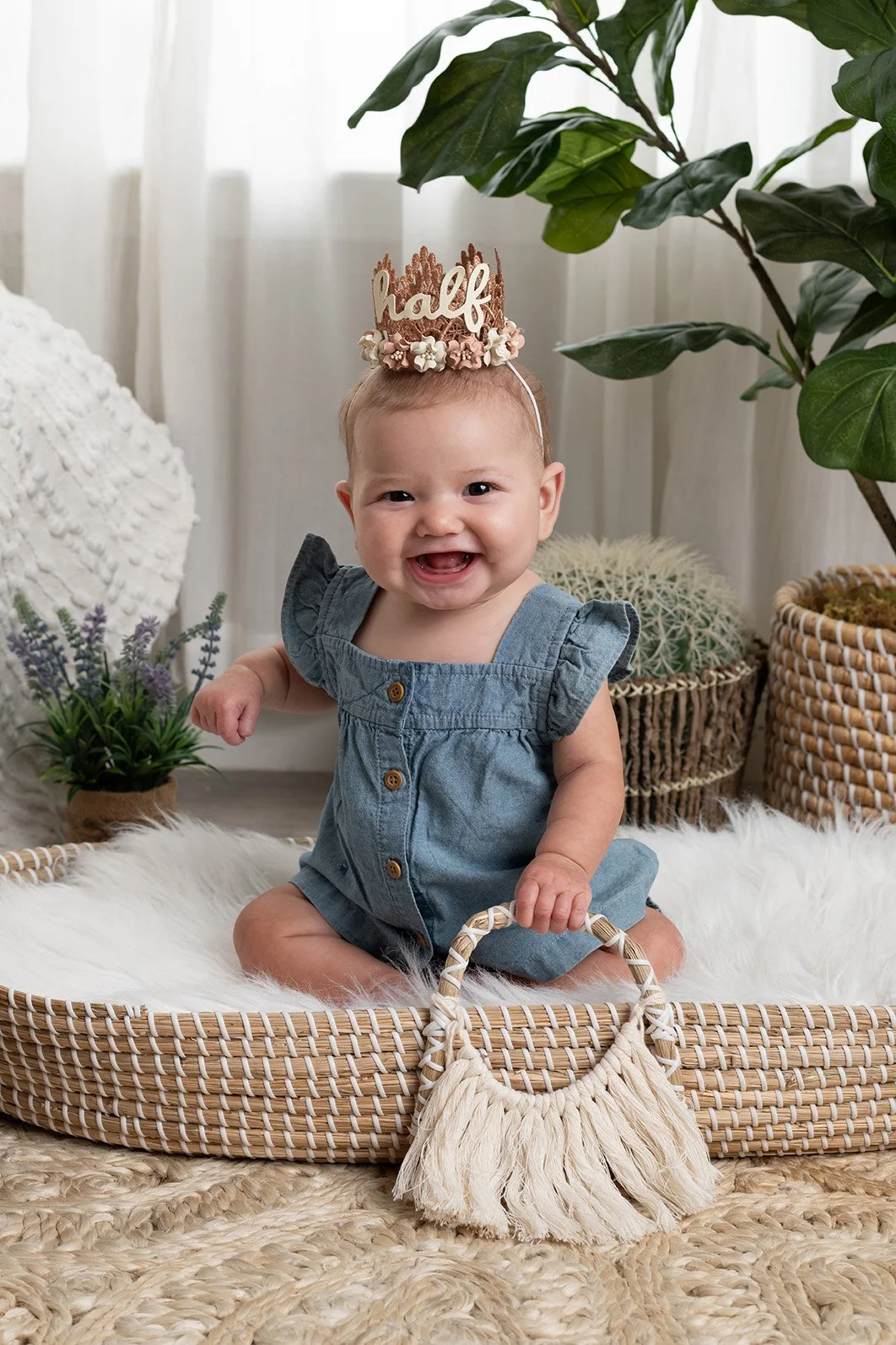 A happy baby girl sitting in a woven basket, wearing a denim dress with ruffled sleeves and a "half" birthday crown, surrounded by greenery and decorative plants.