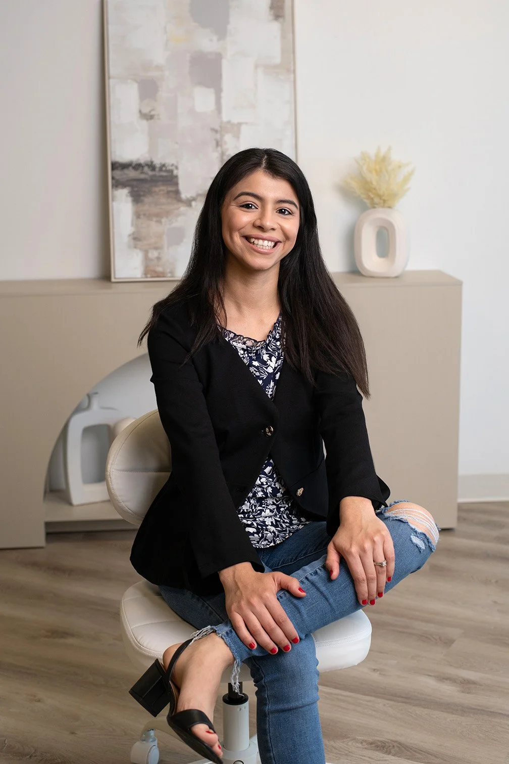 A woman sitting cross-legged on a white chair in an office, smiling at the camera. She has long dark hair, is wearing a black blazer, a patterned blouse, ripped jeans, and black heeled sandals. Behind her is a beige cabinet with a decorative white va