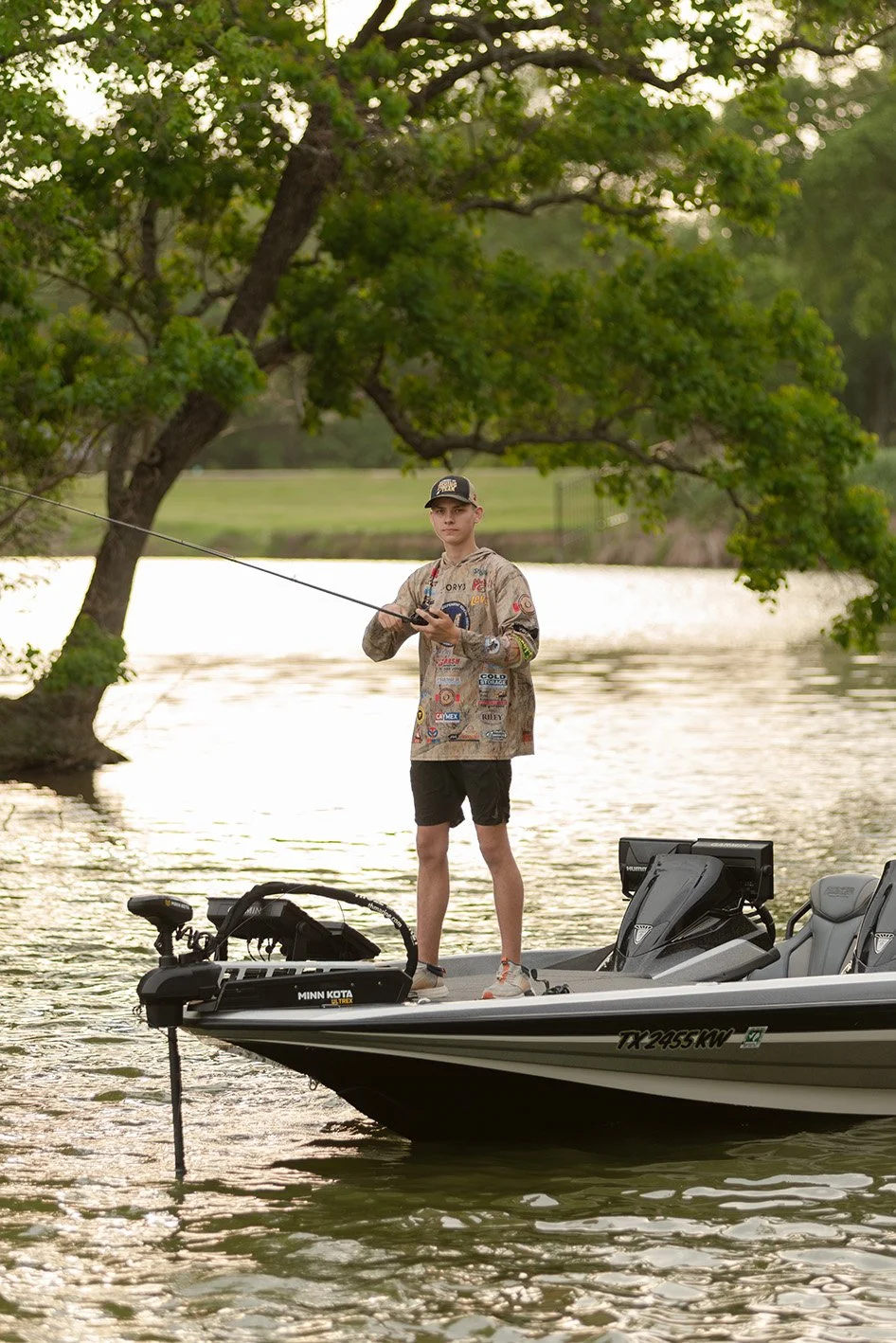 A young man fishing from a boat on a river during daytime, surrounded by trees with green leaves.