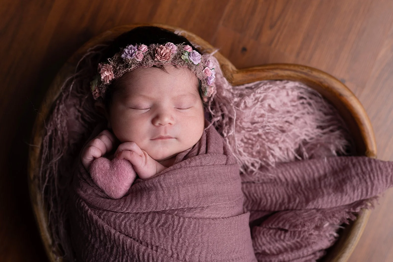 A sleeping baby girl wearing a floral headband, wrapped in a mauve blanket and lying on a fluffy pink blanket inside a wooden bowl.