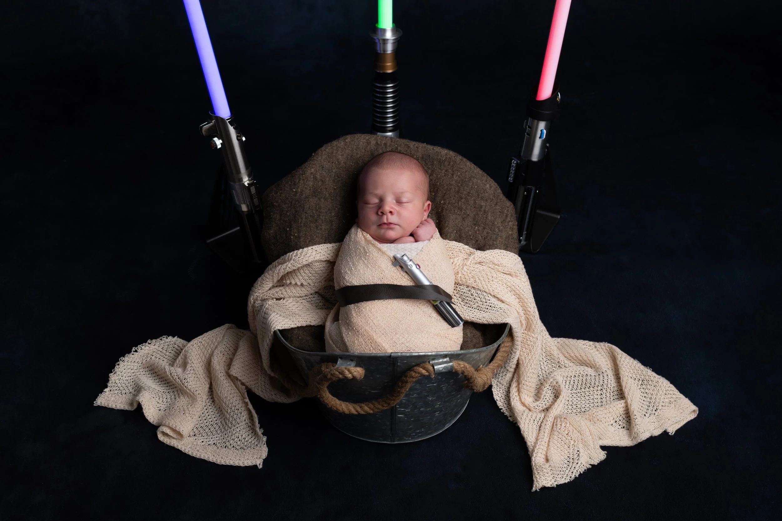 A sleeping baby in a basket with a blanket and straps, surrounded by a dark background and illuminated by colorful lights.