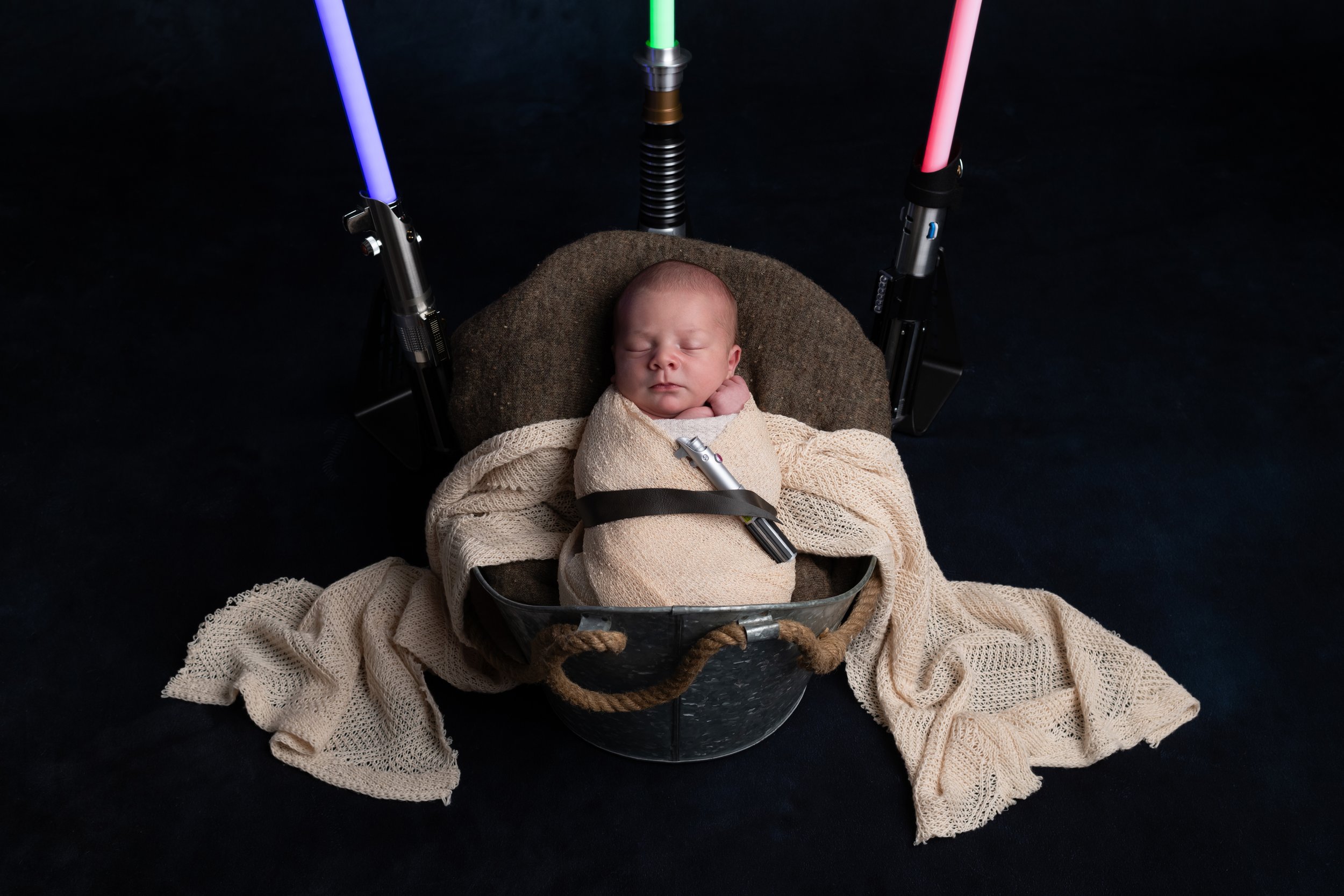 A sleeping baby in a basket with a blanket and straps, surrounded by a dark background and illuminated by colorful lights.