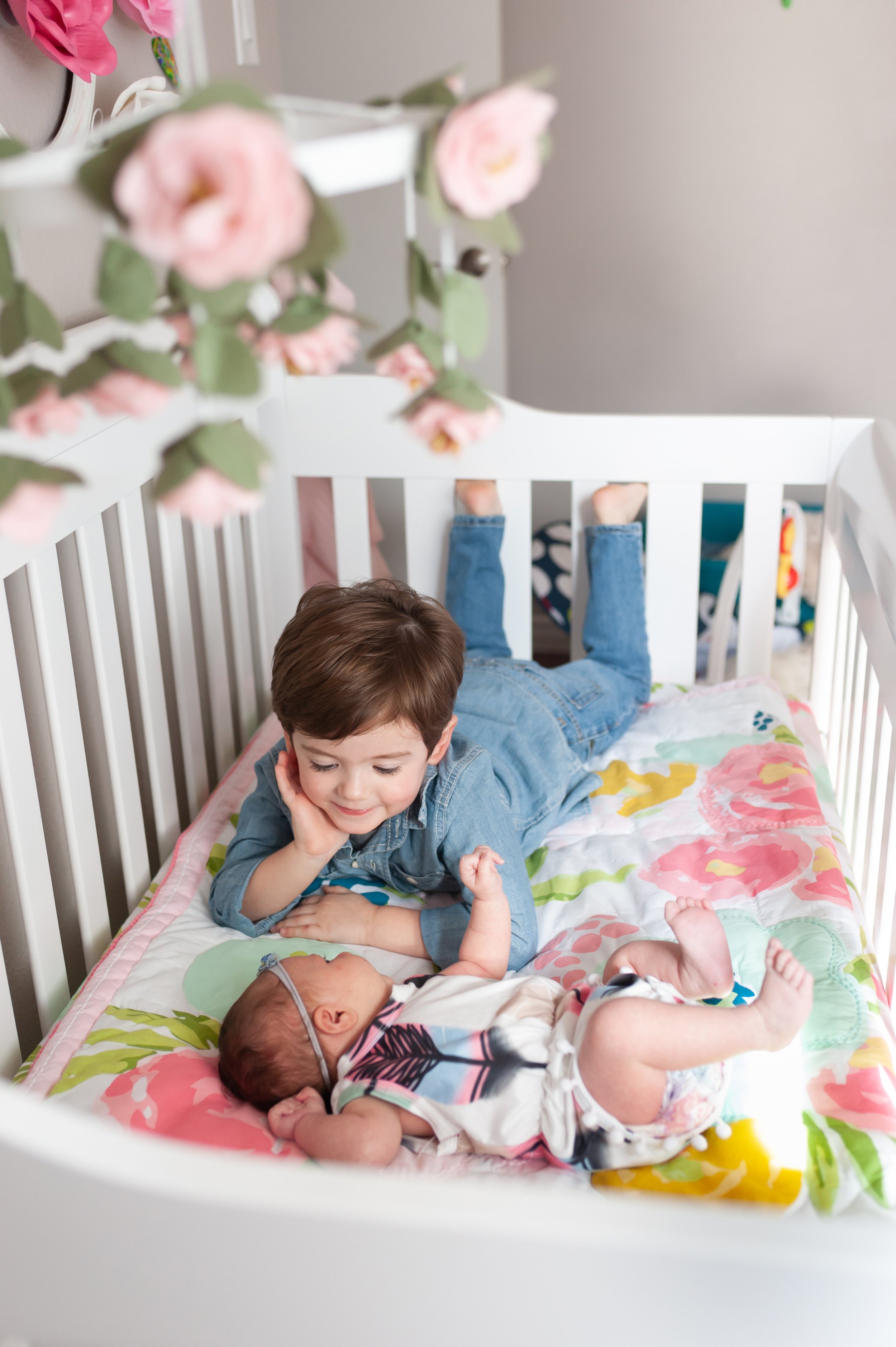 A young boy lying in a white crib, looking at and smiling at a newborn baby girl dressed in pajamas, with a floral blanket covering her. The boy is resting on his arm with a gentle smile, and the scene appears to be in a nursery decorated with flower