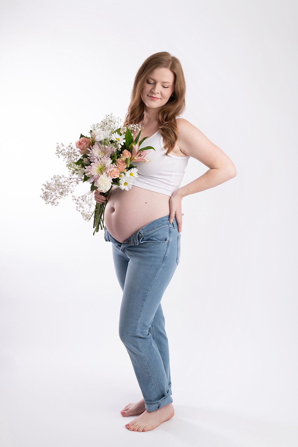 Pregnant woman with red hair wearing a white tank top and jeans holds a bouquet of flowers and stands barefoot against a white background.
