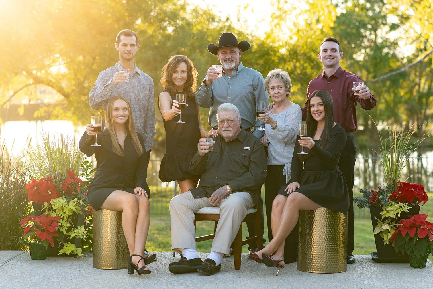 A family gathering outdoors during sunset, with nine people holding wine glasses and celebrating, surrounded by potted plants and holiday decorations.
