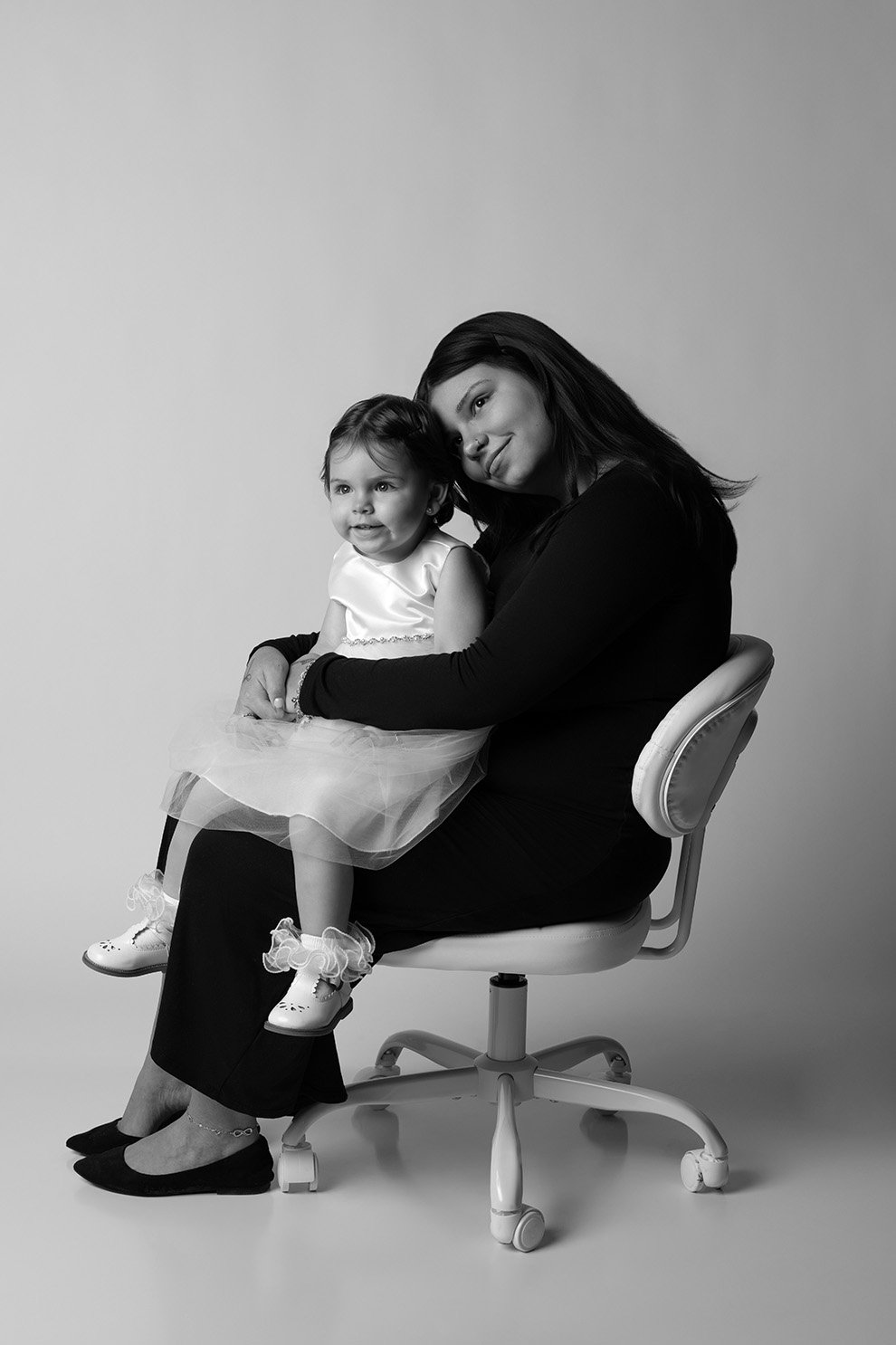 A woman and a young girl sitting together on an office chair, smiling at the camera in a black and white photograph.