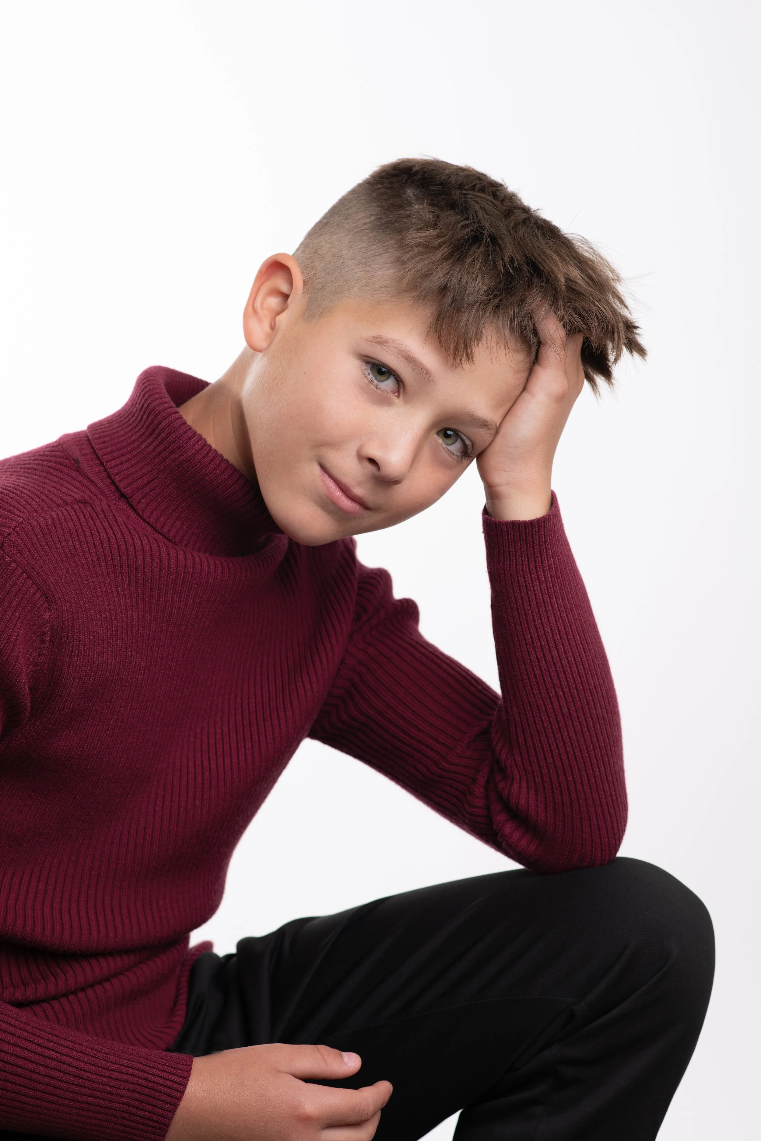 A young boy with short brown hair, wearing a maroon ribbed turtleneck sweater, sitting against a plain white background, resting his head on his hand and looking at the camera.