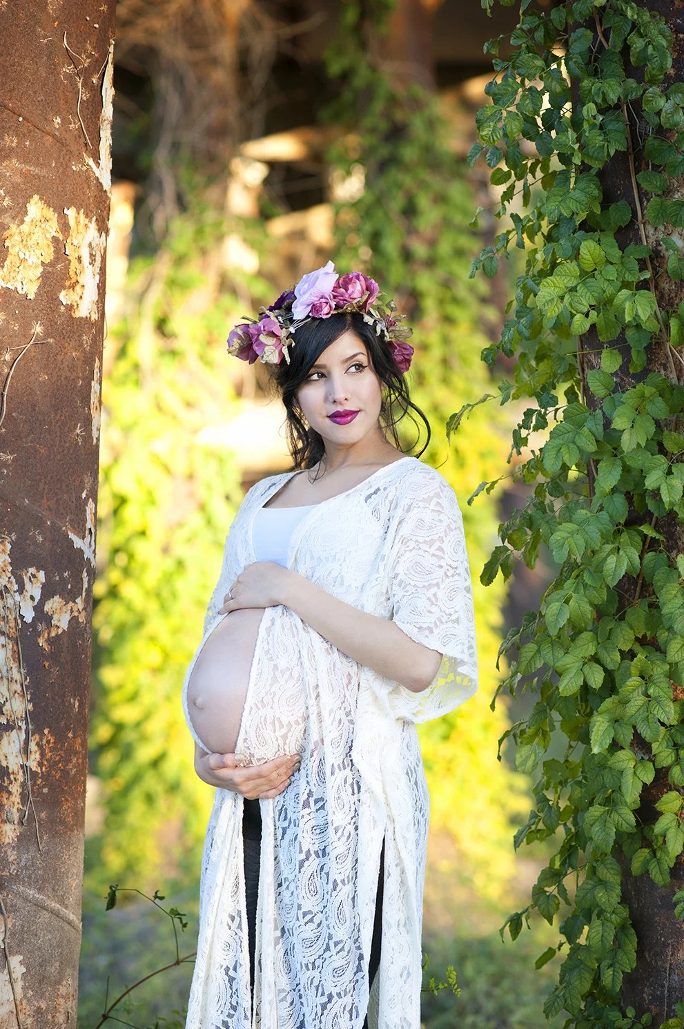 A pregnant woman wearing a white lace dress and a floral crown stands outdoors between two trees covered in green leaves, holding her belly and looking to the side.