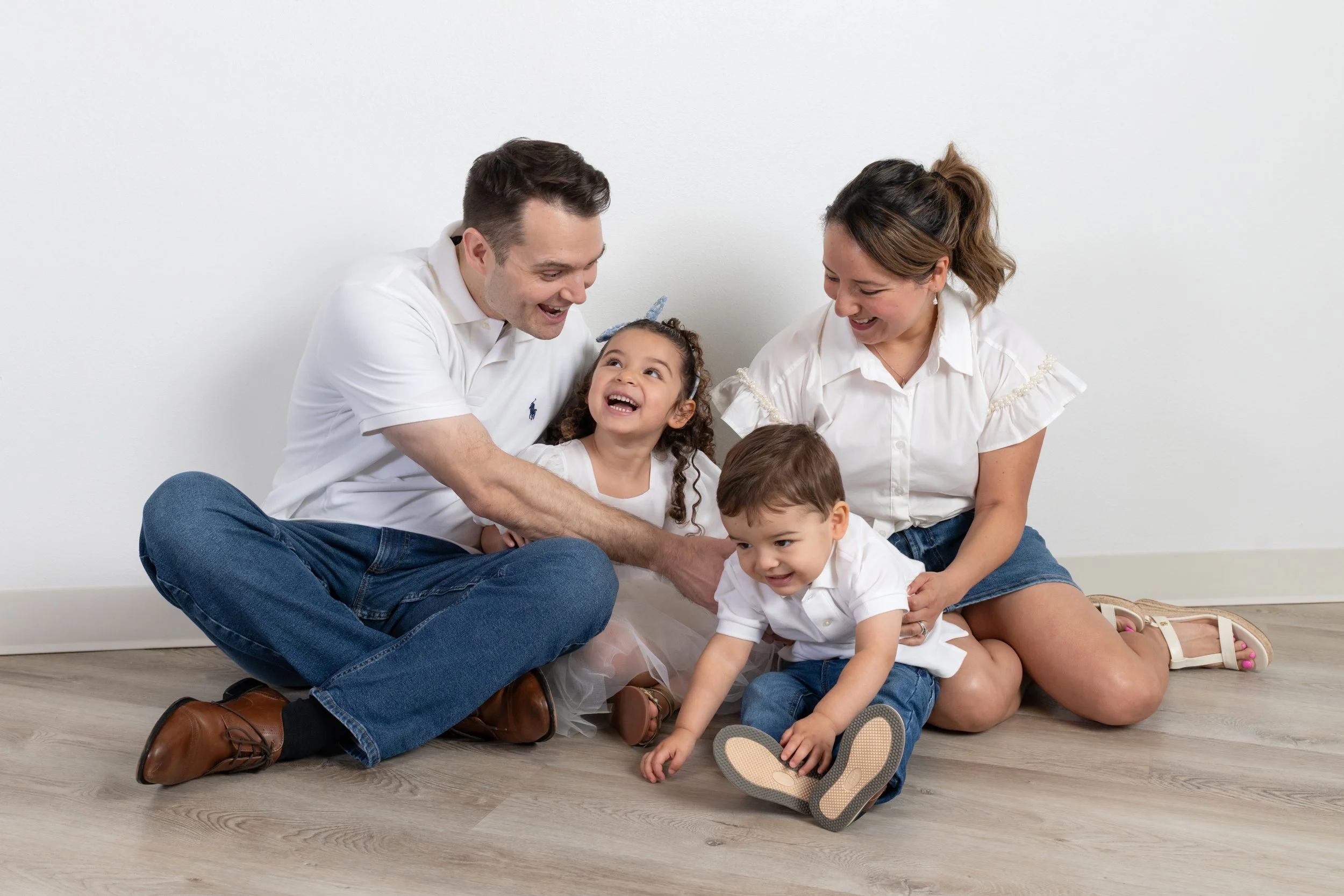 A happy family of four, including two children and their parents, sitting on the floor against a plain white wall, laughing and playing together.