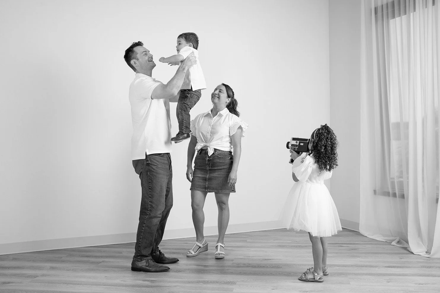 A family of four and a young girl with curly hair taking a picture in a bright room with white walls and curtains, with the family playing and smiling as the father lifts a young boy into the air.