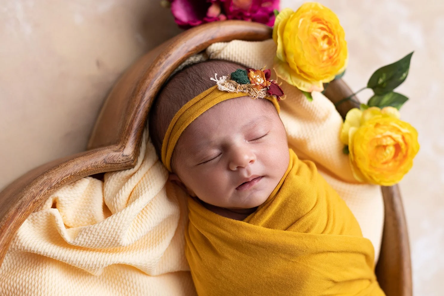A sleeping baby wrapped in a yellow blanket, lying in a wooden cradle, decorated with yellow and pink flowers and a floral headband.