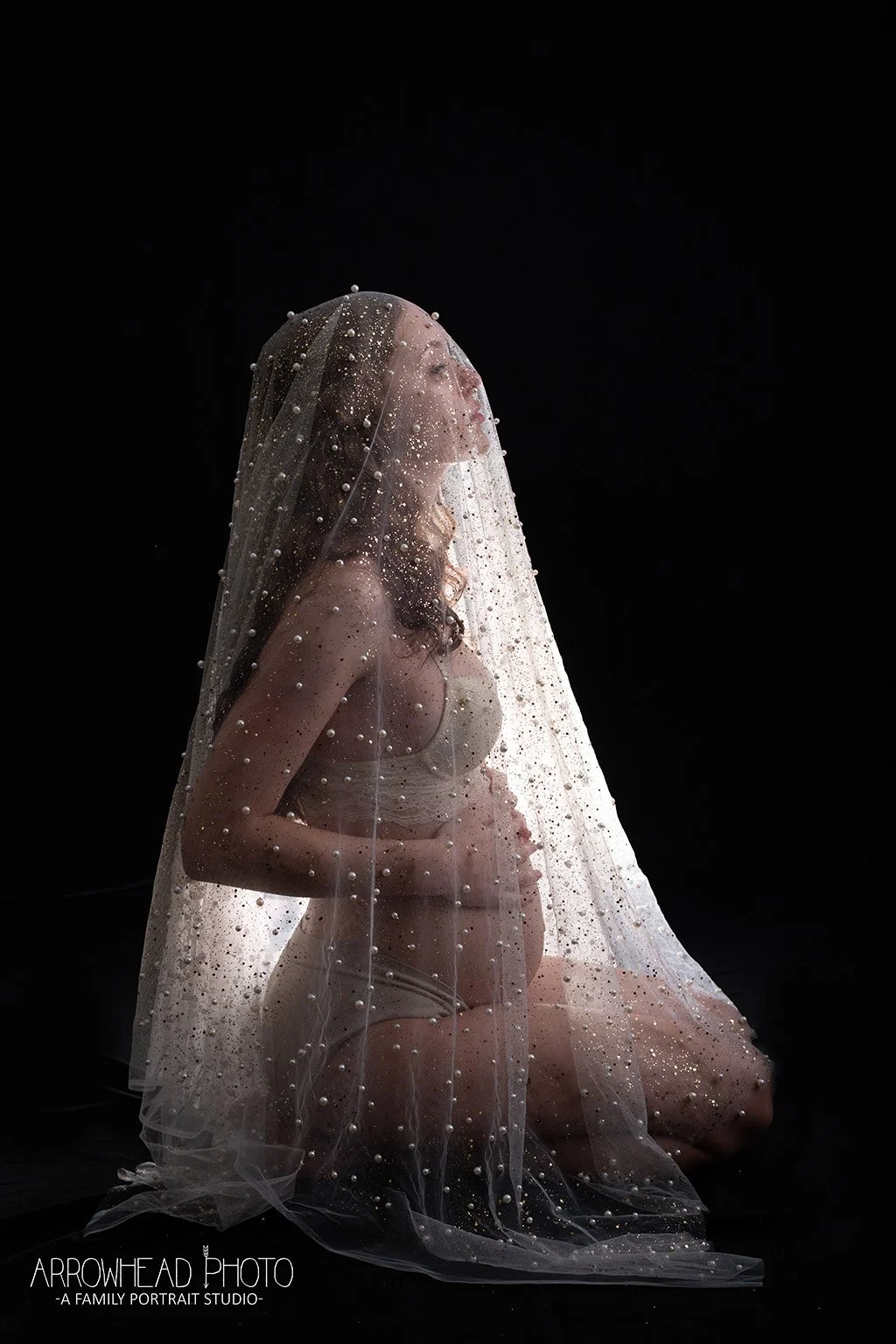 A pregnant woman sitting on her knees in profile, draped with a sheer veil decorated with pearls, against a black background.