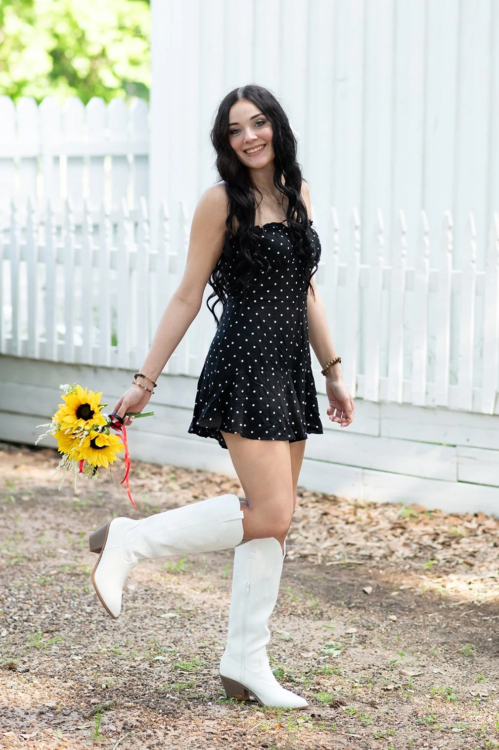 A woman with long dark hair, wearing a black dress with white polka dots and white knee-high boots, is holding a bouquet of sunflowers and small white flowers and smiling.
