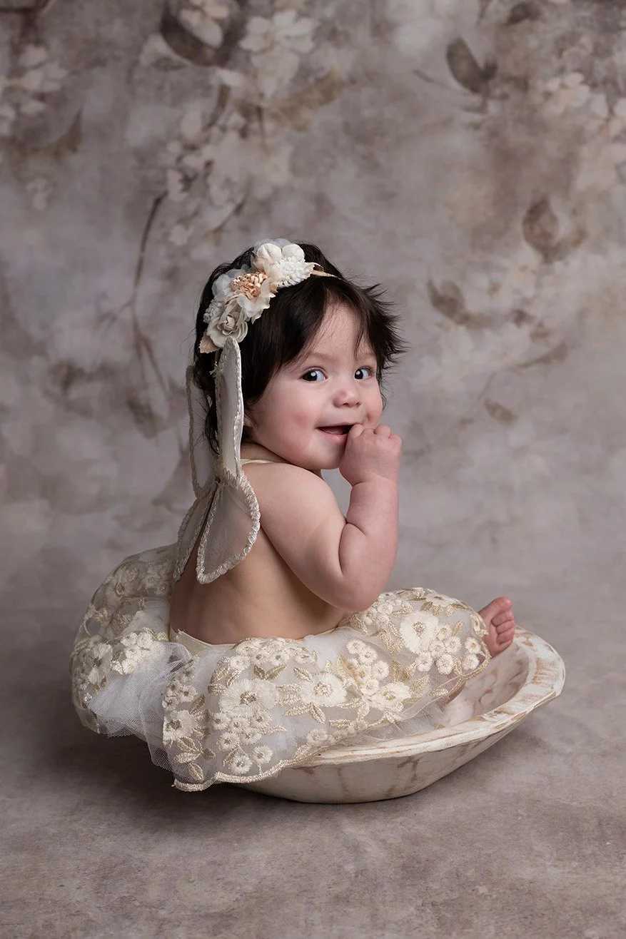 Adorable baby girl with dark hair, wearing a flower headband, sitting in a cream-colored floral dress inside a shallow bowl, against a soft, floral background, smiling and playing with her hand.