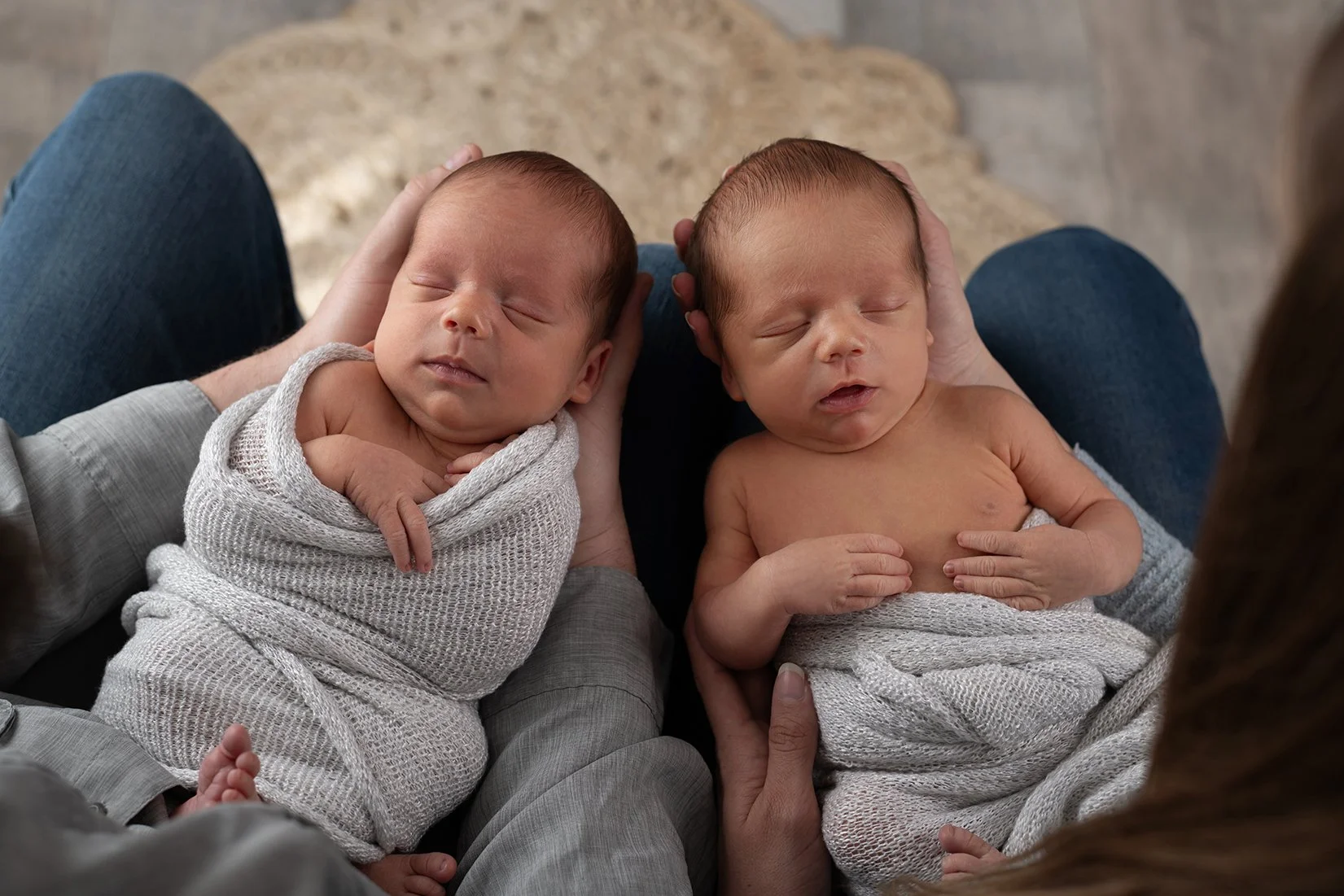 Two sleeping newborn babies, wrapped in gray blankets, are held in an adult's arms while sitting on the adult's lap. The babies have closed eyes and peaceful expressions.