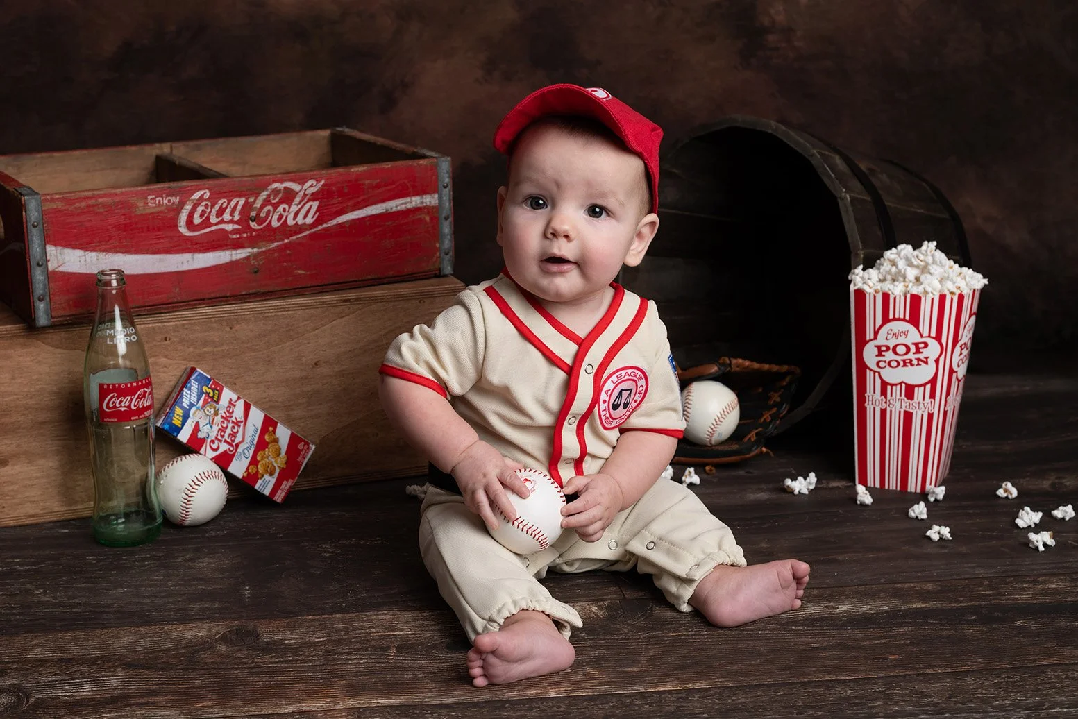 Baby sitting on wooden floor in baseball uniform, surrounded by popcorn, soda, baseball, and a box of crackers, with an open baseball glove and a wooden box in the background.