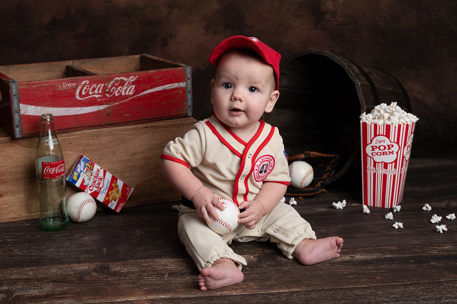 Baby sitting on wooden floor in baseball uniform, surrounded by popcorn, soda, baseball, and a box of crackers, with an open baseball glove and a wooden box in the background.