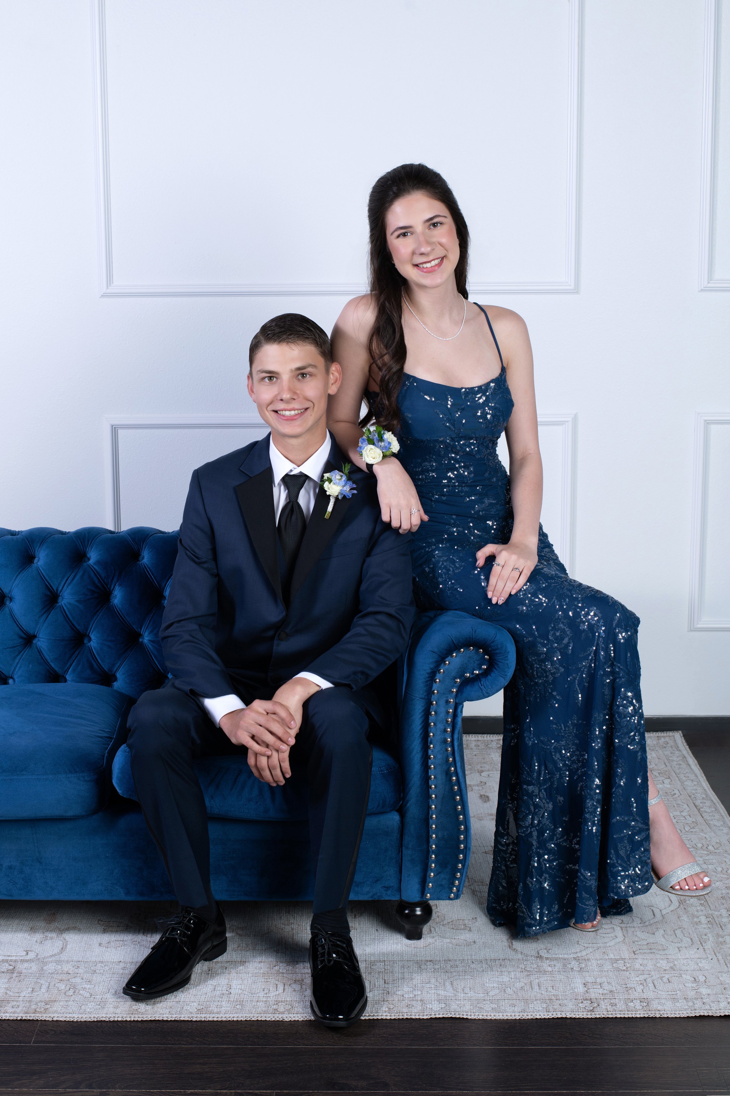 A young man in a dark blue tuxedo and a young woman in a long, navy blue sequin gown pose together in a formal setting. The man is seated on a blue tufted sofa, smiling with hands clasped, and the woman is standing next to him, resting her hand on hi
