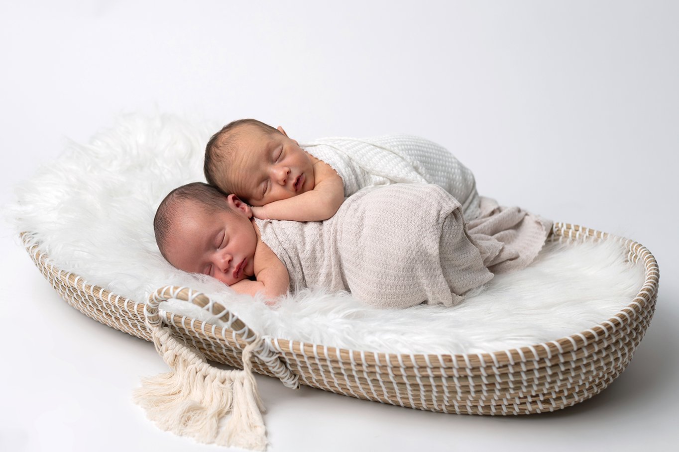 Two sleeping newborn babies lying on a soft white blanket inside a woven basket.