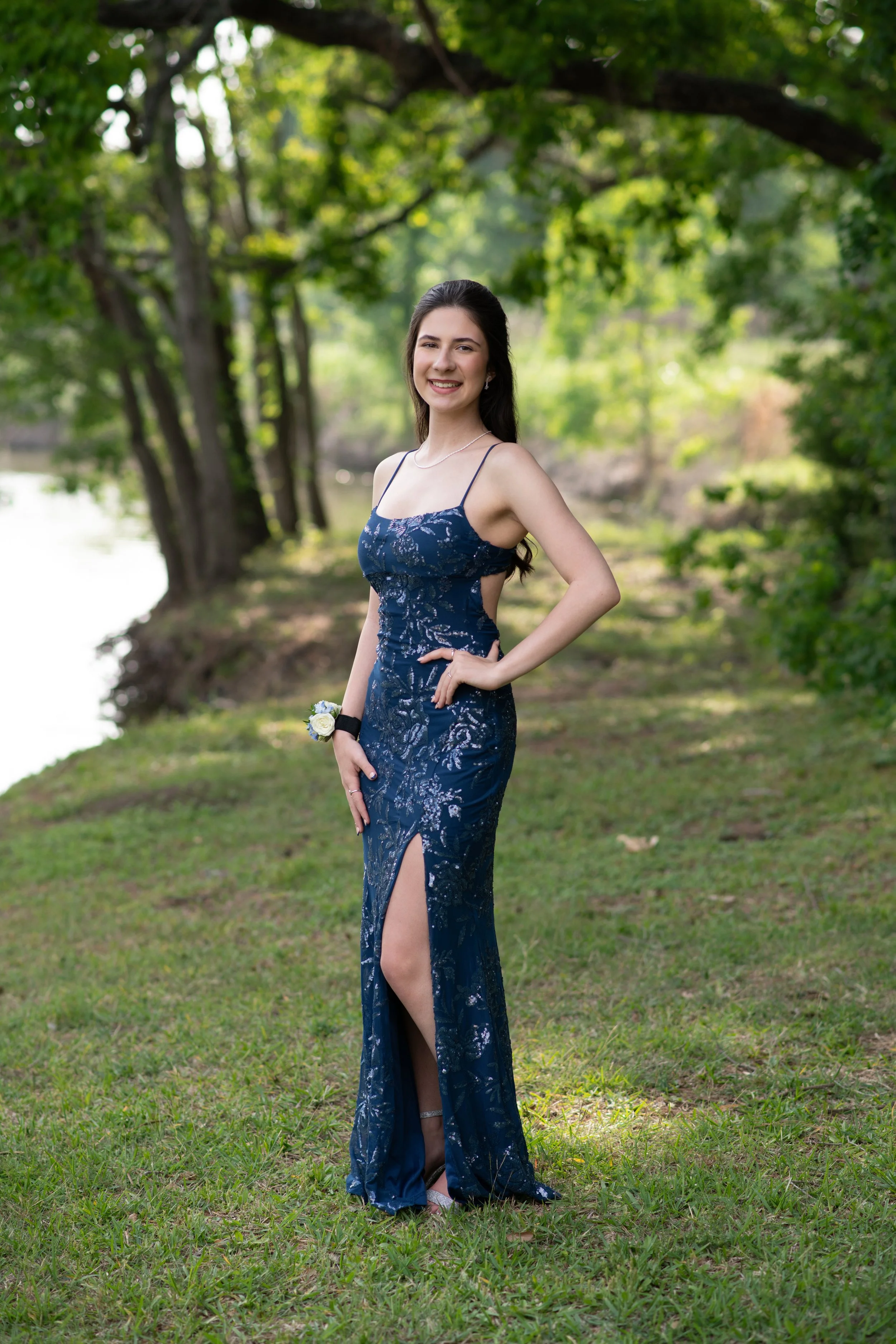 A young woman in a dark blue, strapless, sequined gown with a thigh-high slit, standing outdoors on grass by a wooded area, smiling with one hand on her hip and holding a small bouquet in her other hand.
