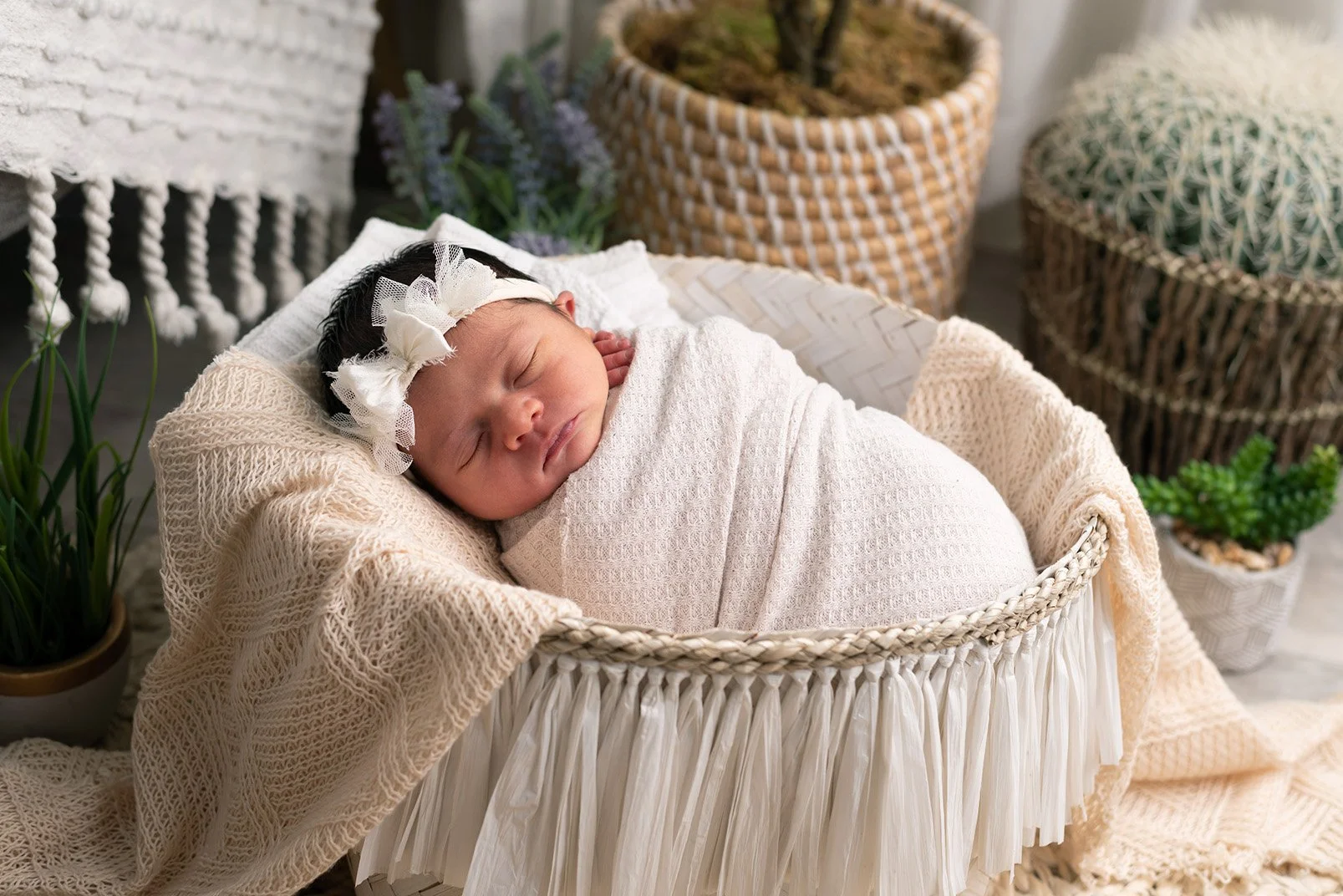 A sleeping baby wrapped in a cream blanket, lying in a woven bassinet in a decorated indoor setting with plants and woven baskets in the background.