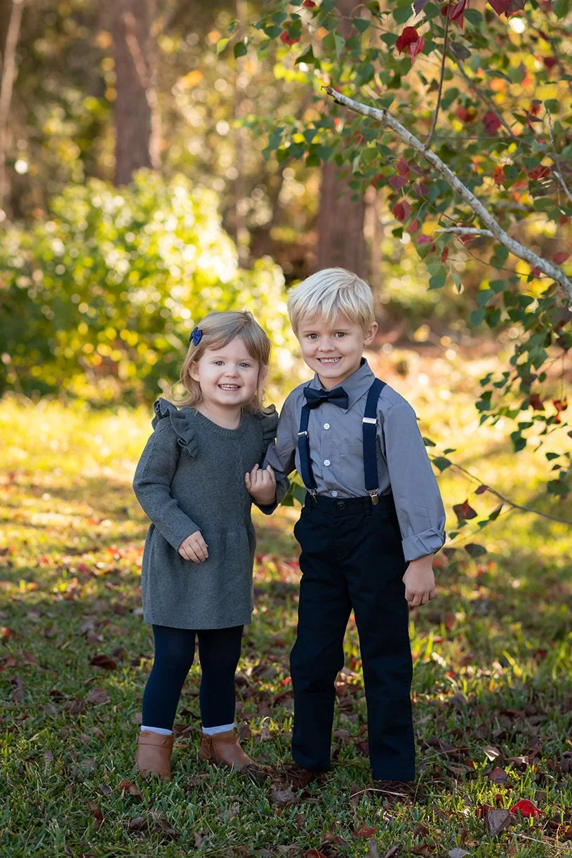 A young girl and boy standing outdoors in a wooded area on a sunny day, smiling and holding hands. The girl is wearing a gray dress, blue tights, and brown boots, with a blue hair clip. The boy is dressed in a gray shirt, black pants, suspenders, and