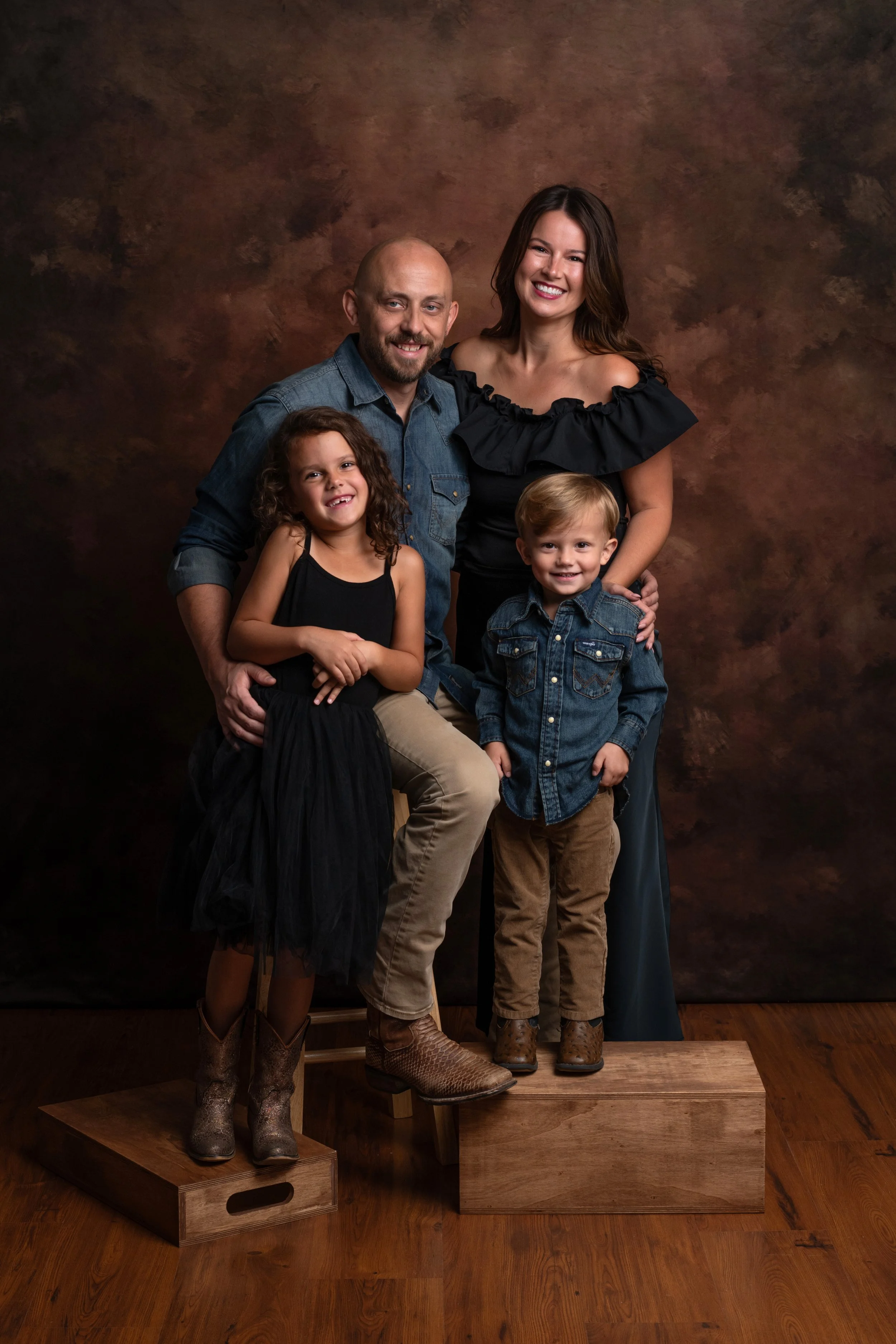 Family of four, two adults and two children, posing together on wooden boxes with a brown textured background. All are smiling, dressed in casual clothing, with the woman wearing an off-shoulder top. The children are wearing denim and black dresses. 