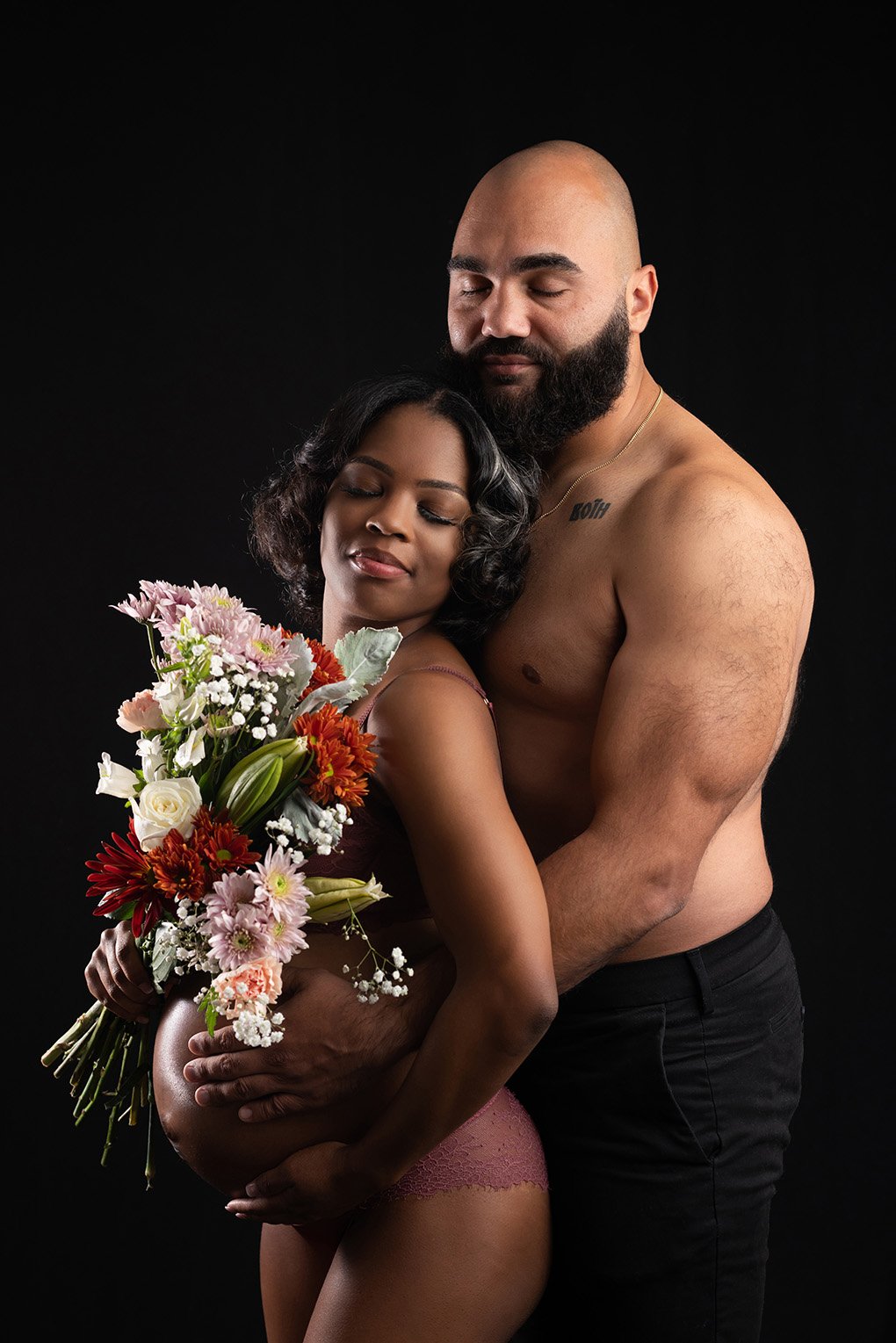 A pregnant woman with dark skin and black hair holding a colorful bouquet of flowers, embracing a shirtless man with a beard and light skin against a black background.