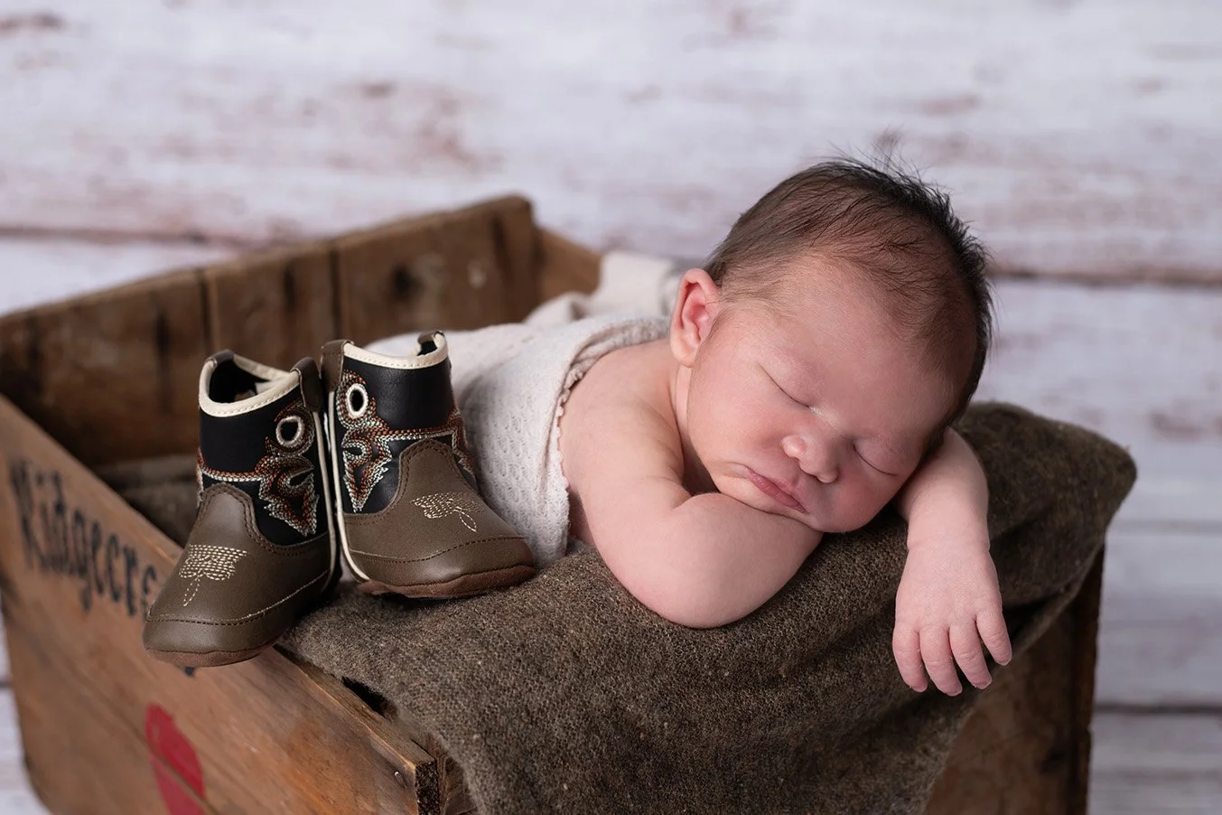 A sleeping baby lying on a brown cushion in a rustic wooden box with a pair of brown hiking boots nearby.