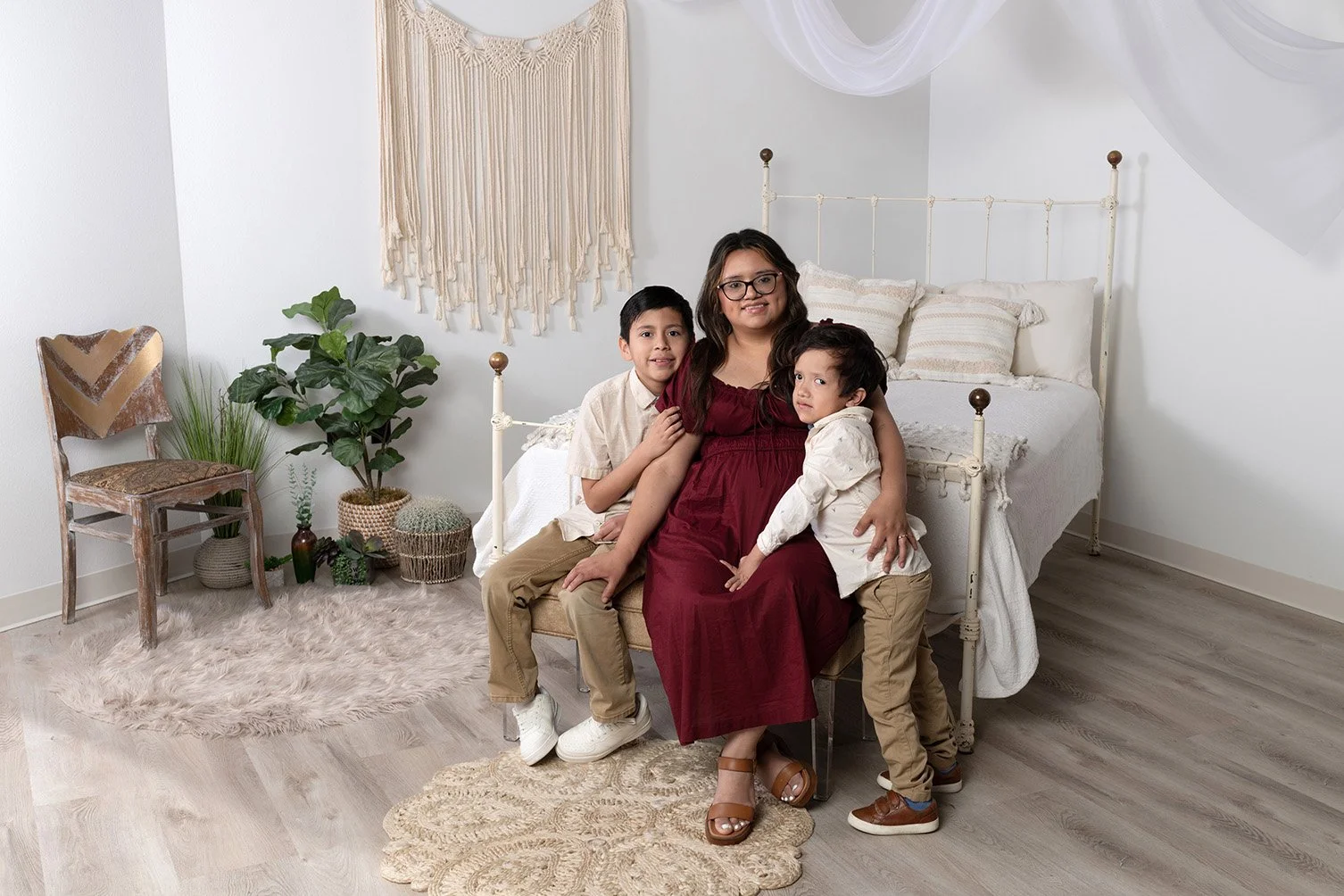 A woman with two young boys sitting on a bench in a bedroom with a white bed, green plants, a wooden chair, and beige decor, smiling at the camera.