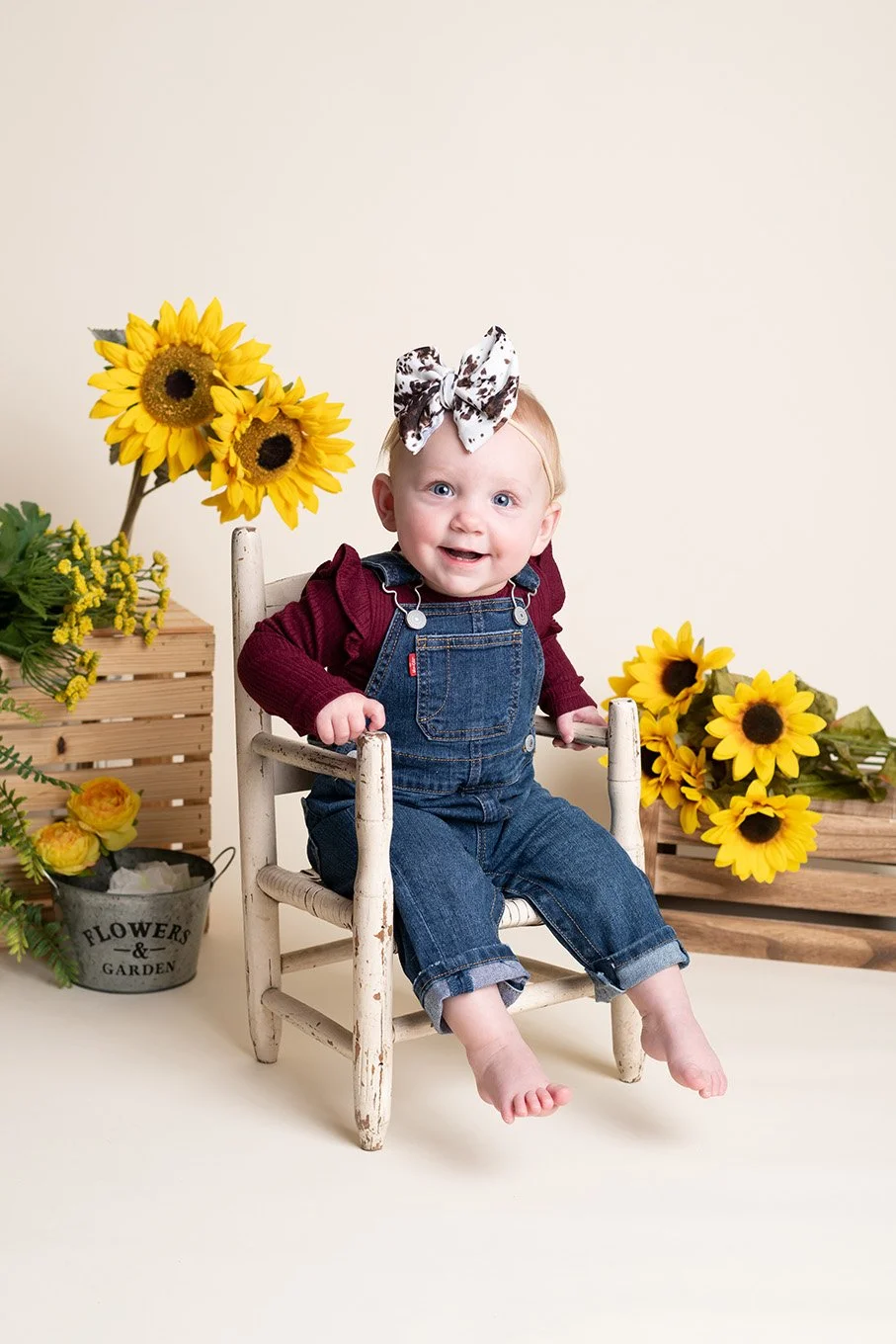 A smiling baby girl with a sunflower headband, wearing denim overalls and a maroon long-sleeve shirt, sitting on a white wooden chair surrounded by sunflowers and garden flowers.