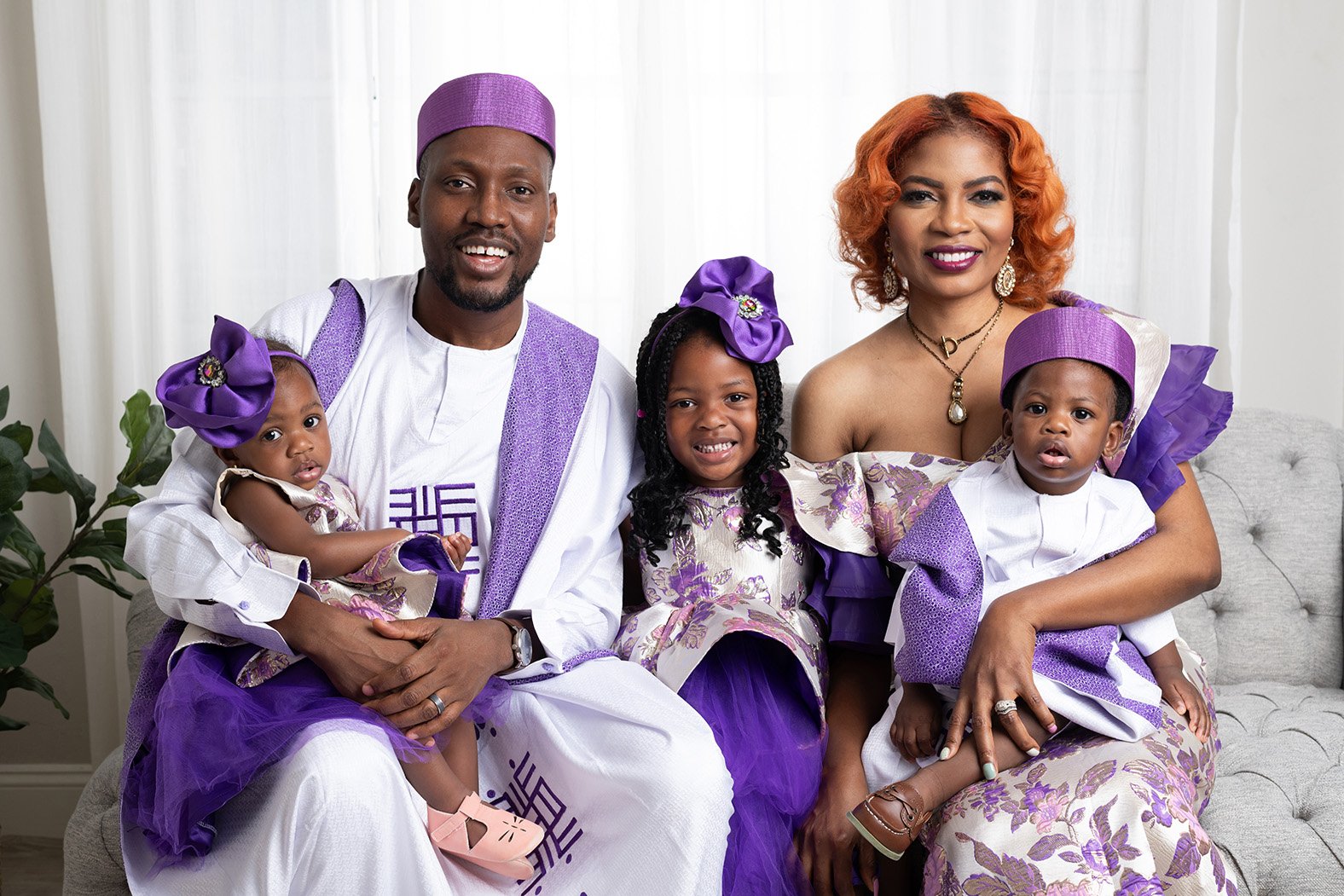 A family of five sitting on a sofa, dressed in purple and white traditional Nigerian attire. The father and two children are wearing matching outfits, while the mother and daughter are dressed elegantly. The family is smiling, showcasing a joyful mom