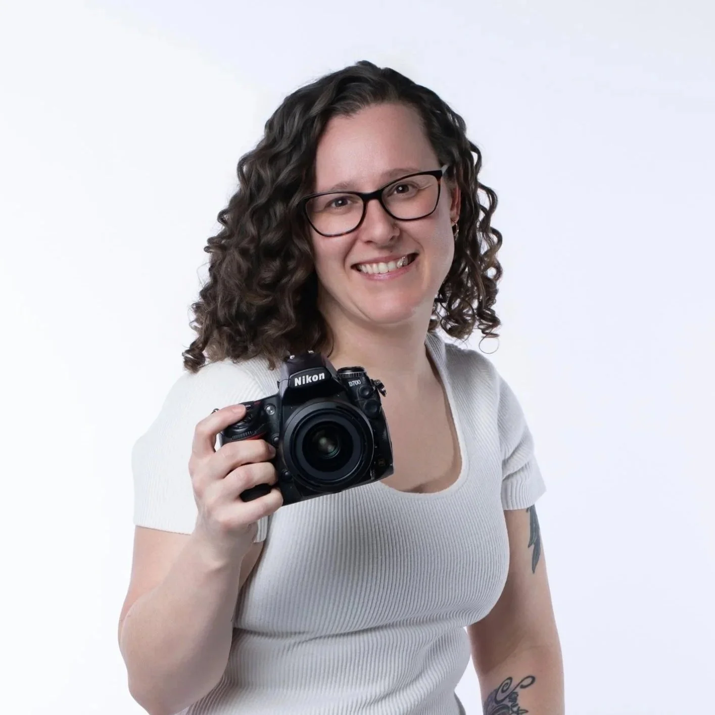 A woman with curly brown hair, glasses, and a white short-sleeve shirt holds a Nikon DSLR camera, smiling against a plain white background.