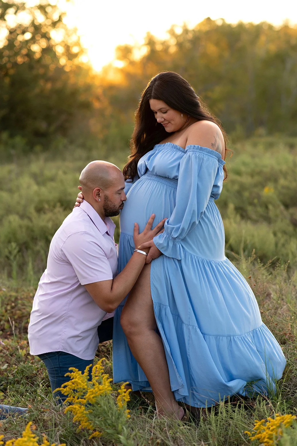 A pregnant woman in a flowing blue dress with an off-the-shoulder neckline stands in a grassy field at sunset, with a man kneeling in front of her, kissing her baby bump. The man has a bald head and is wearing a light pink shirt. The woman has long dark hair and is smiling softly. Yellow wildflowers are in the grass, and trees are blurred in the background.